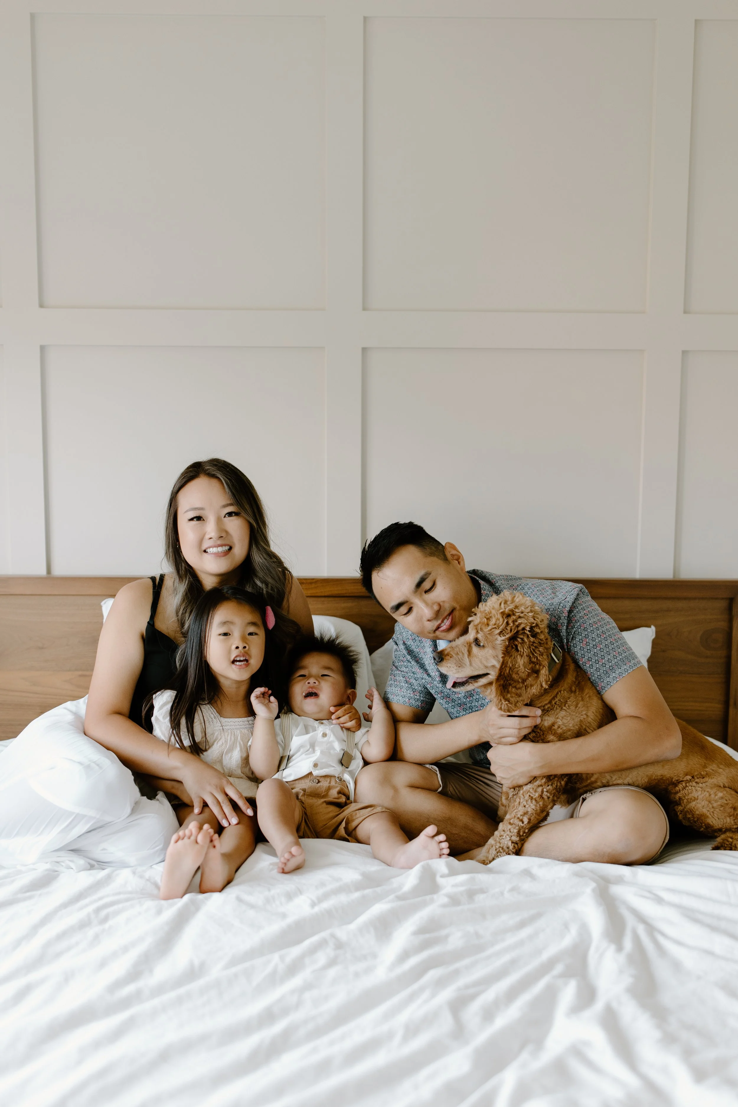Happy family sitting on a bed with two young kids and an adult male holding a dog, in a bedroom with white paneled walls.