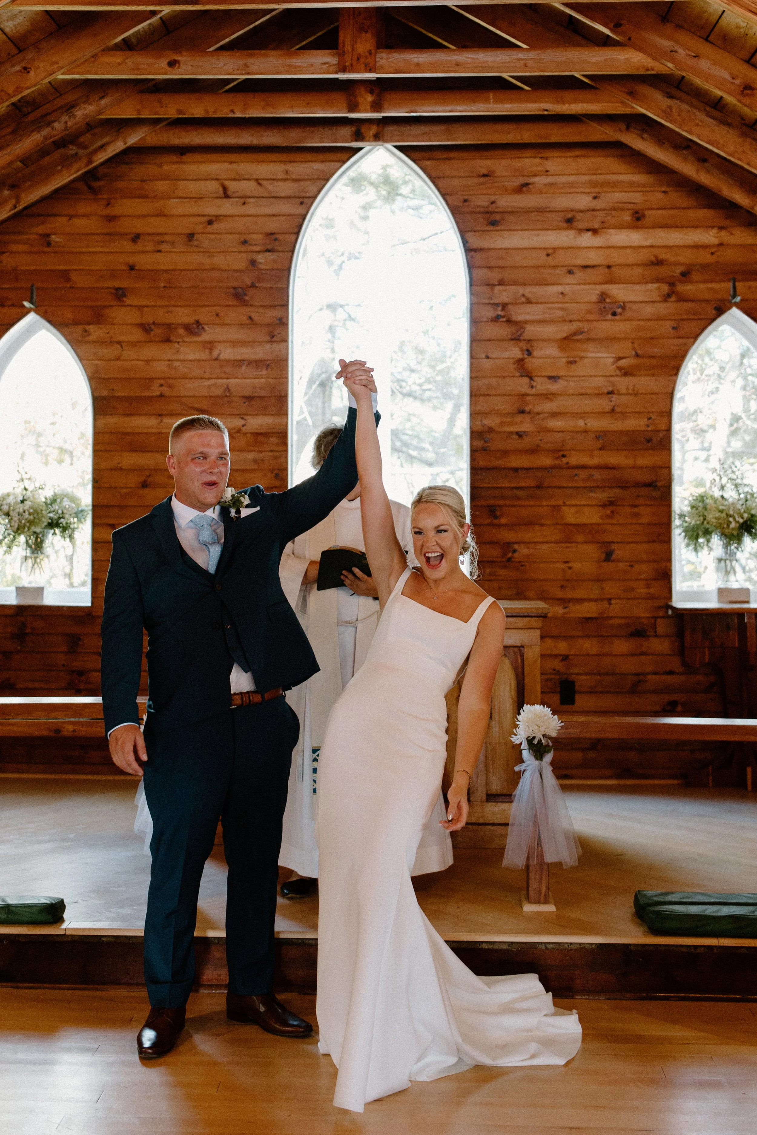 A newly married couple celebrates their wedding inside a wooden church, with the groom holding the bride's hand up high, both smiling joyfully.