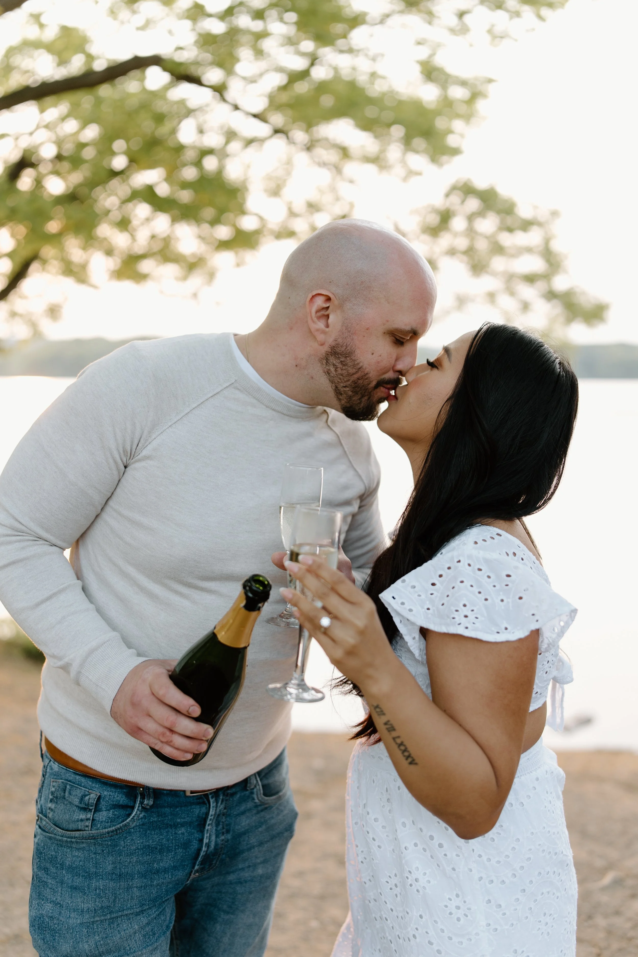 A couple sharing a kiss near a lake, holding champagne and glasses, with trees and water in the background during sunset.