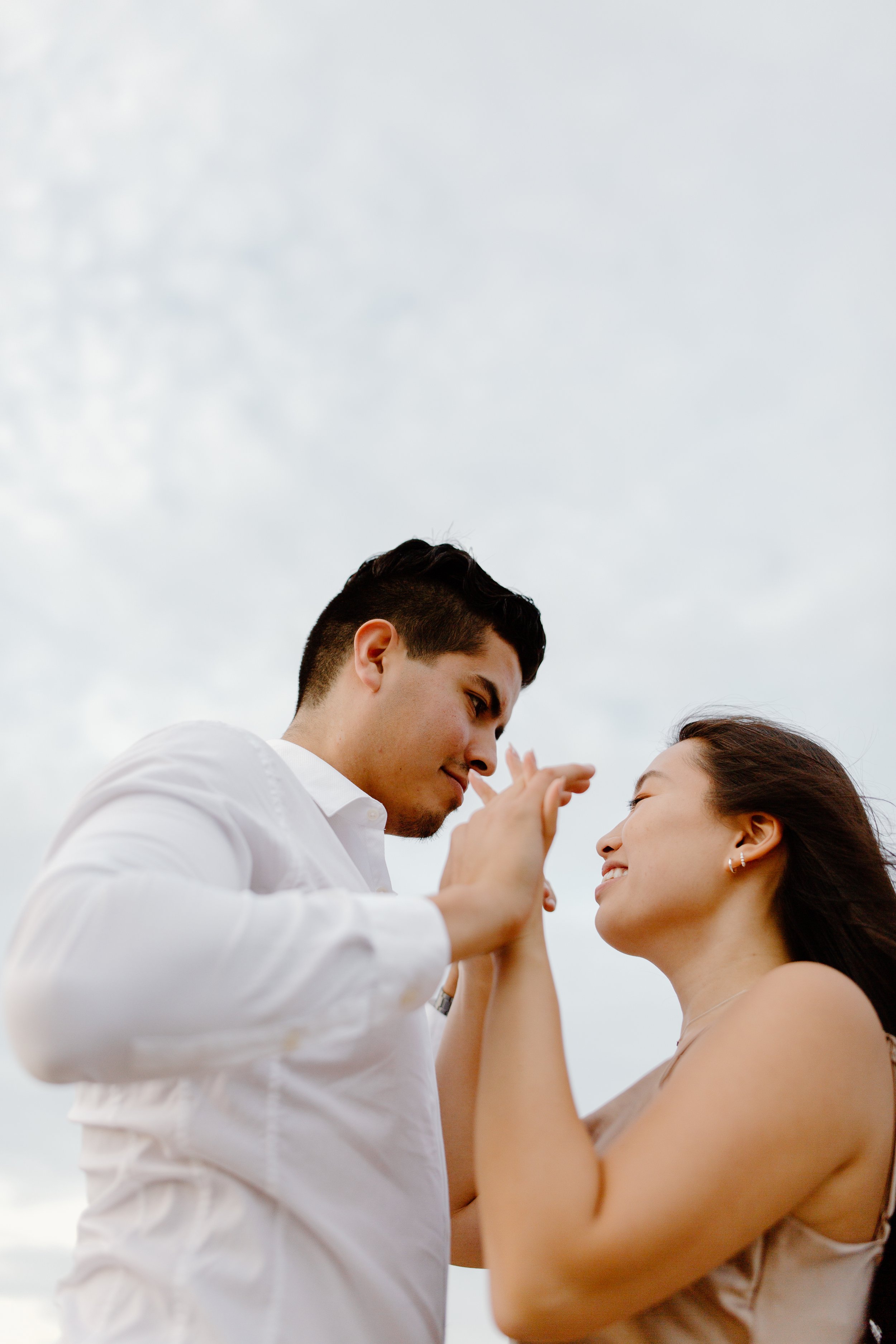 A young man and woman dance closely outdoors, smiling and touching hands, against a cloudy sky.