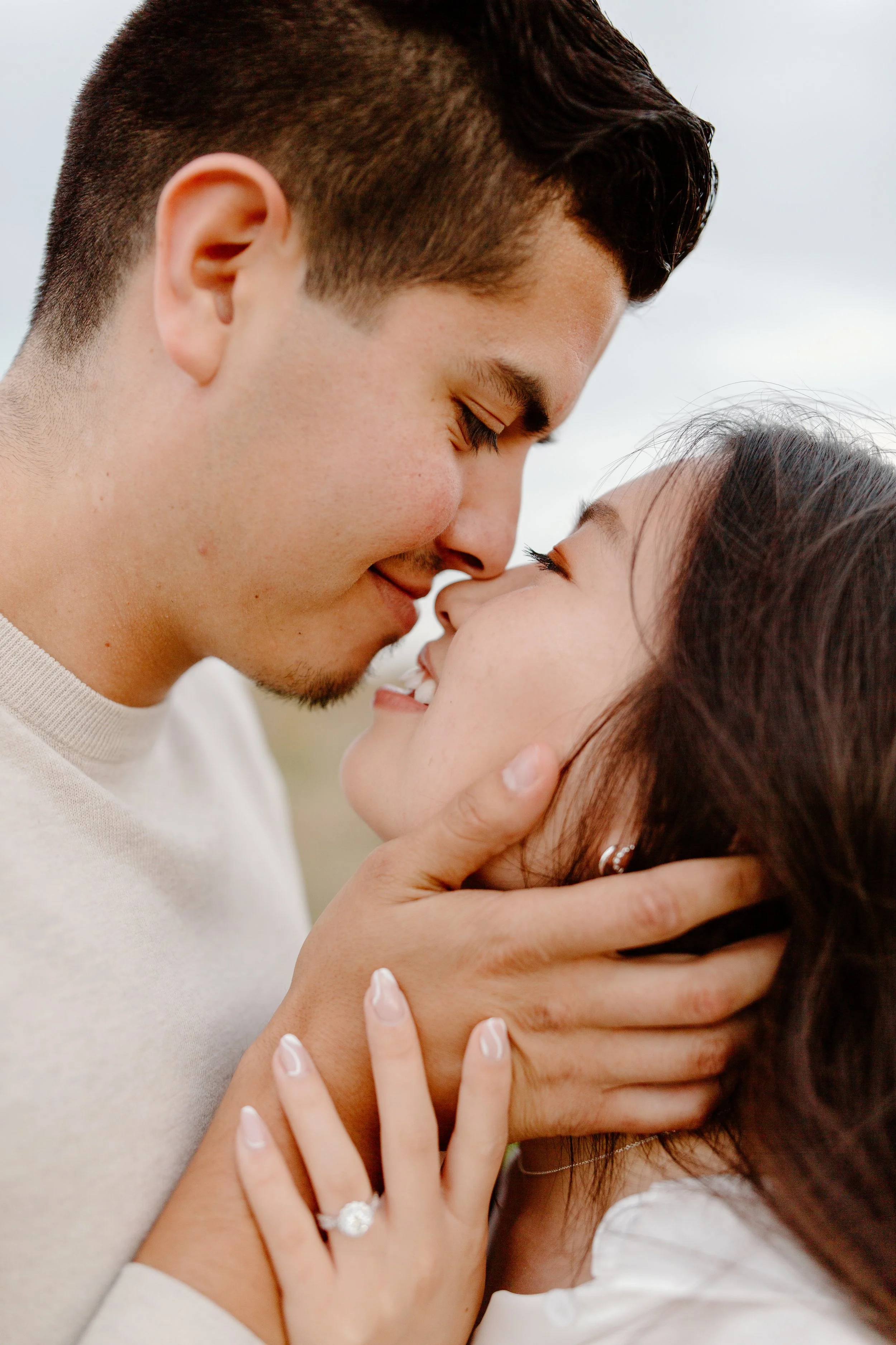 A close-up of a couple with their foreheads touching, smiling, and embracing outdoors. The woman wears a ring and earrings, and the man has short dark hair and a slight beard.