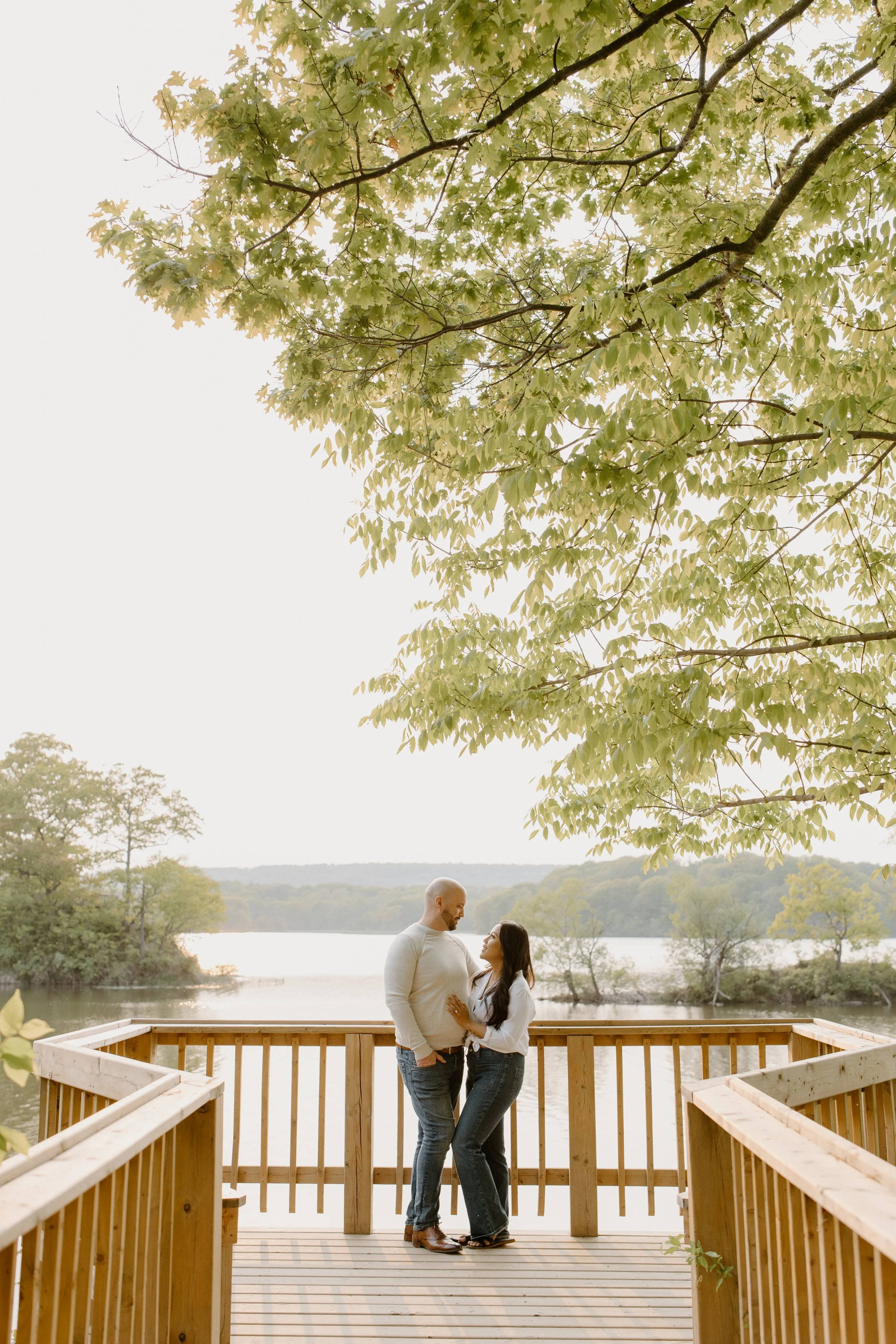 A couple stands on a wooden dock by a lake, gazing at each other romantically, with trees and a calm lake in the background.