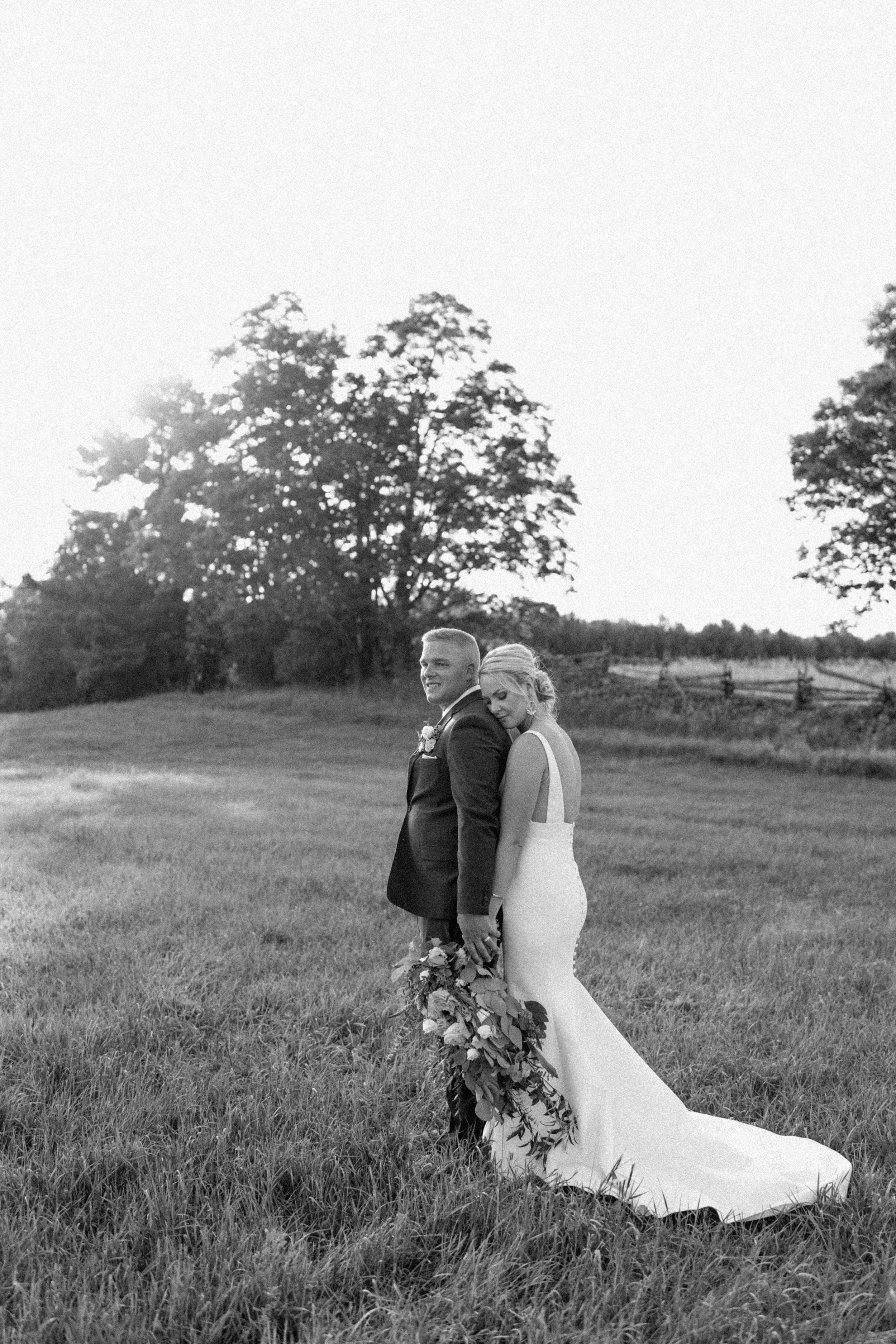 Black and white photo of a bride and groom standing in a grassy field with trees in the background, the bride leaning her head on the groom's shoulder, holding a large bouquet of flowers.