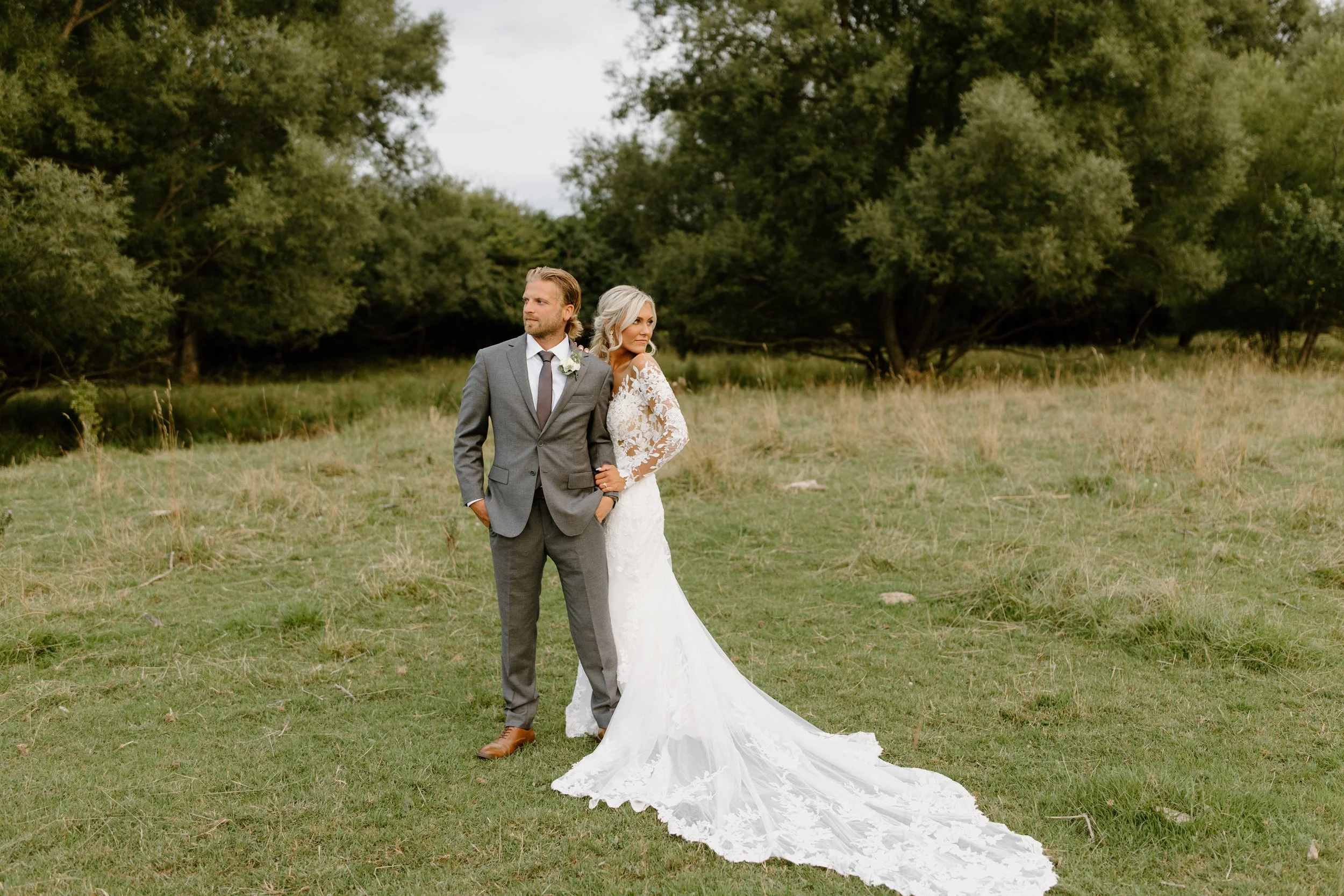 A bride and groom standing together outdoors on a grassy field with trees in the background. The bride wears a white lace wedding dress with train, and the groom wears a grey suit with a white shirt and tie.