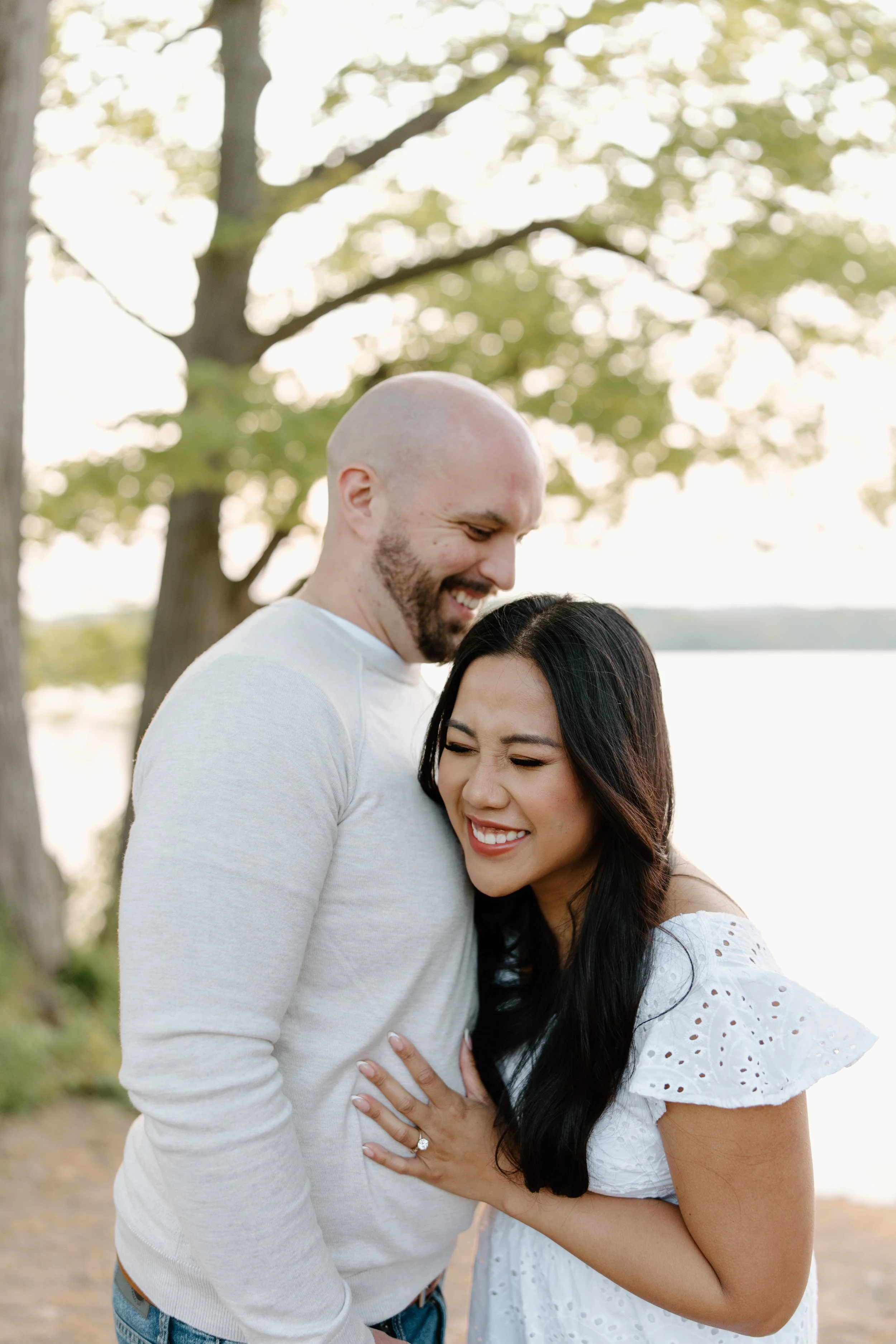 A couple sharing a joyful moment outdoors by a lake, with trees and water in the background, smiling and embracing each other.