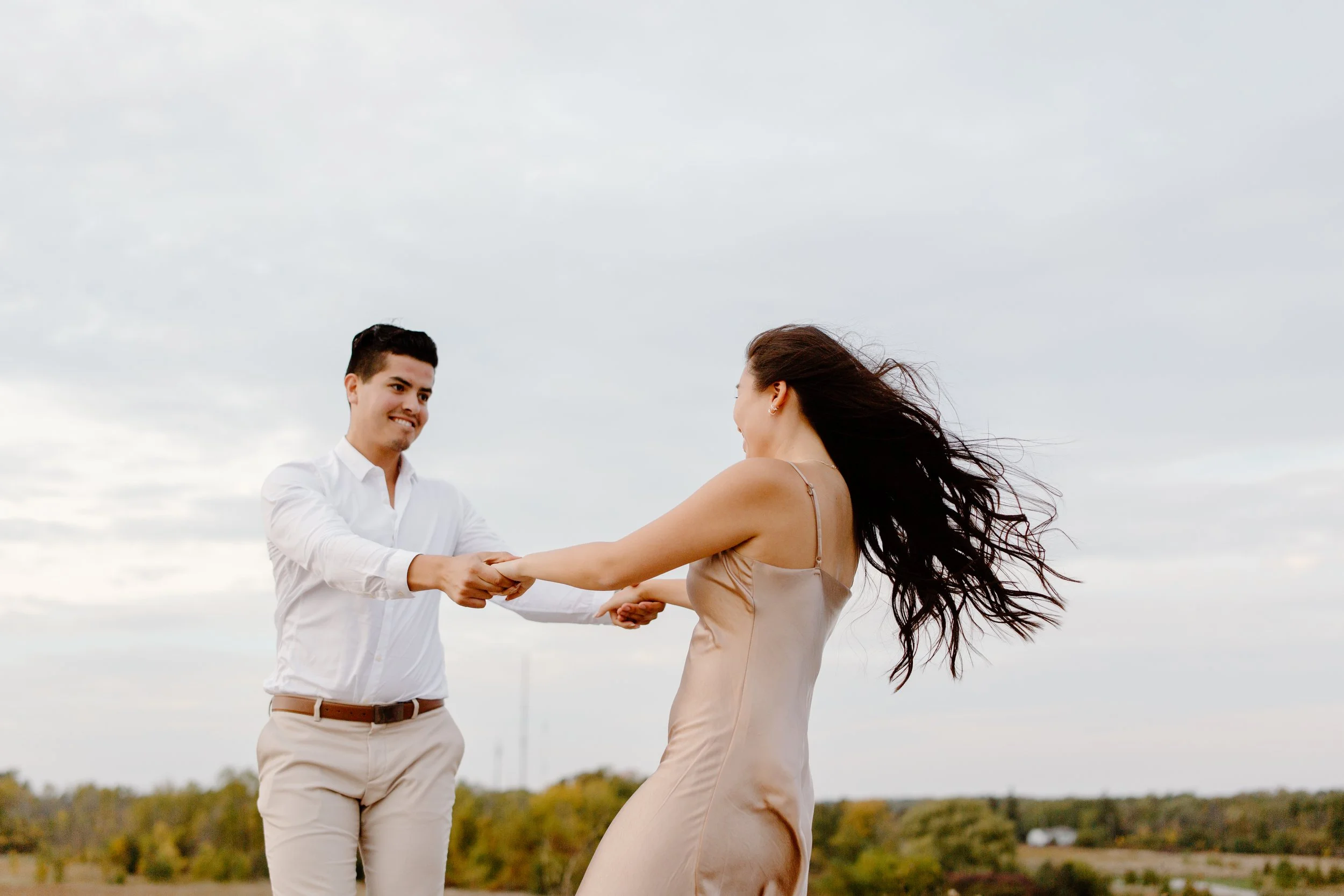 A man and woman holding hands and dancing outdoors on a cloudy day, with trees and fields in the background.