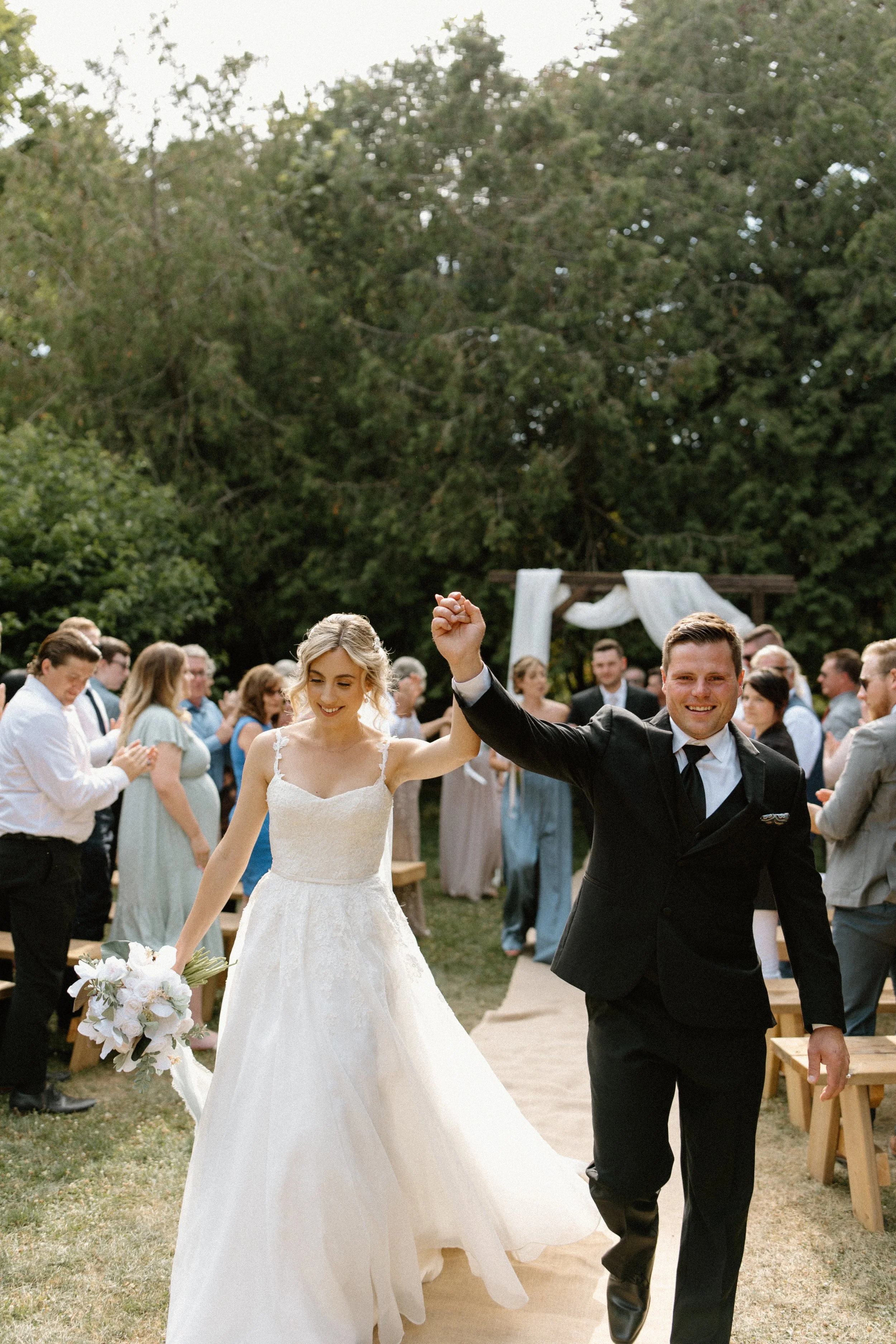 A newlywed couple holding hands and smiling as they walk down the aisle outdoors during their wedding celebration, with guests clapping and a decorated arch in the background.