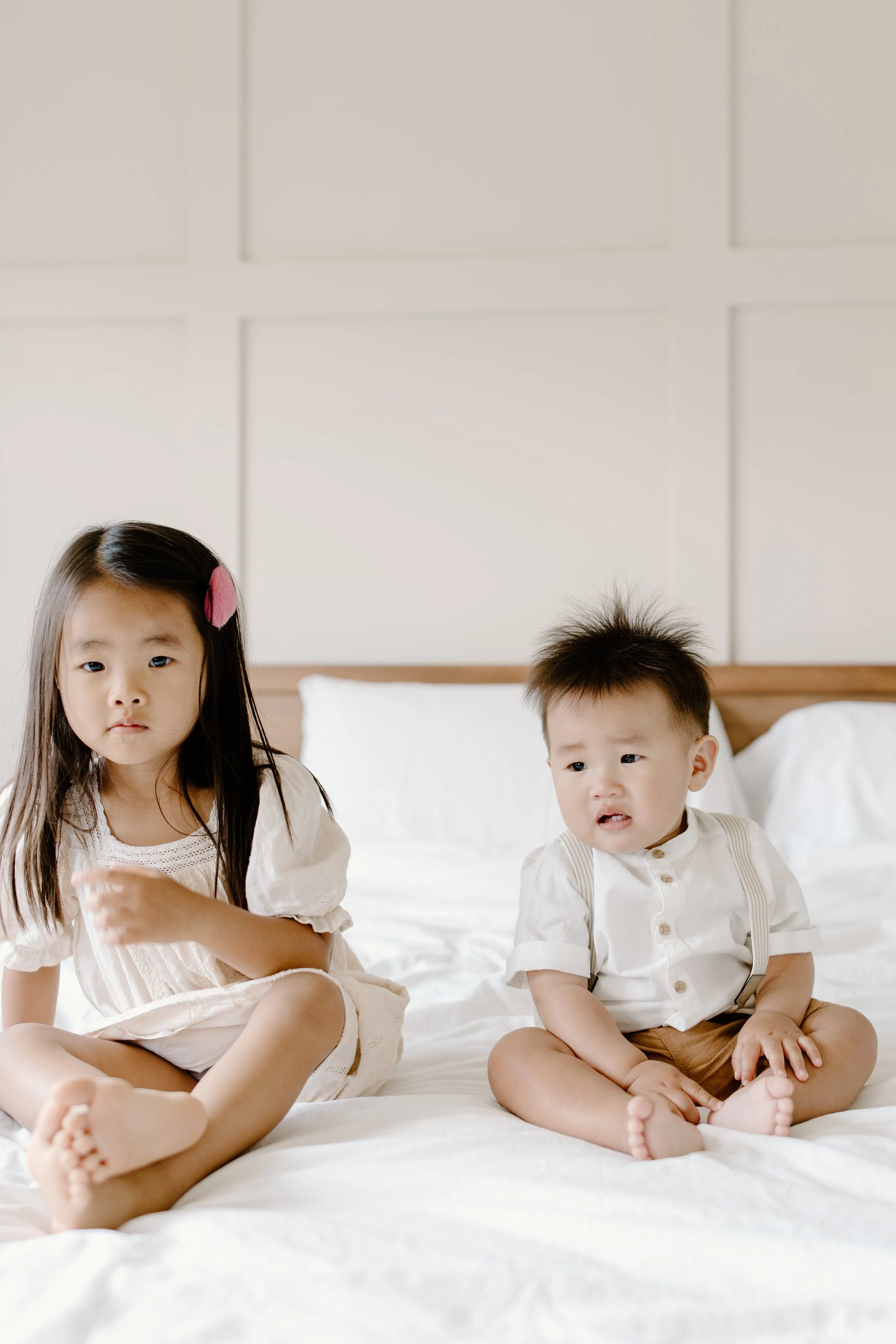 Two young children sitting on a bed, looking confused or sad, with a plain wall background.