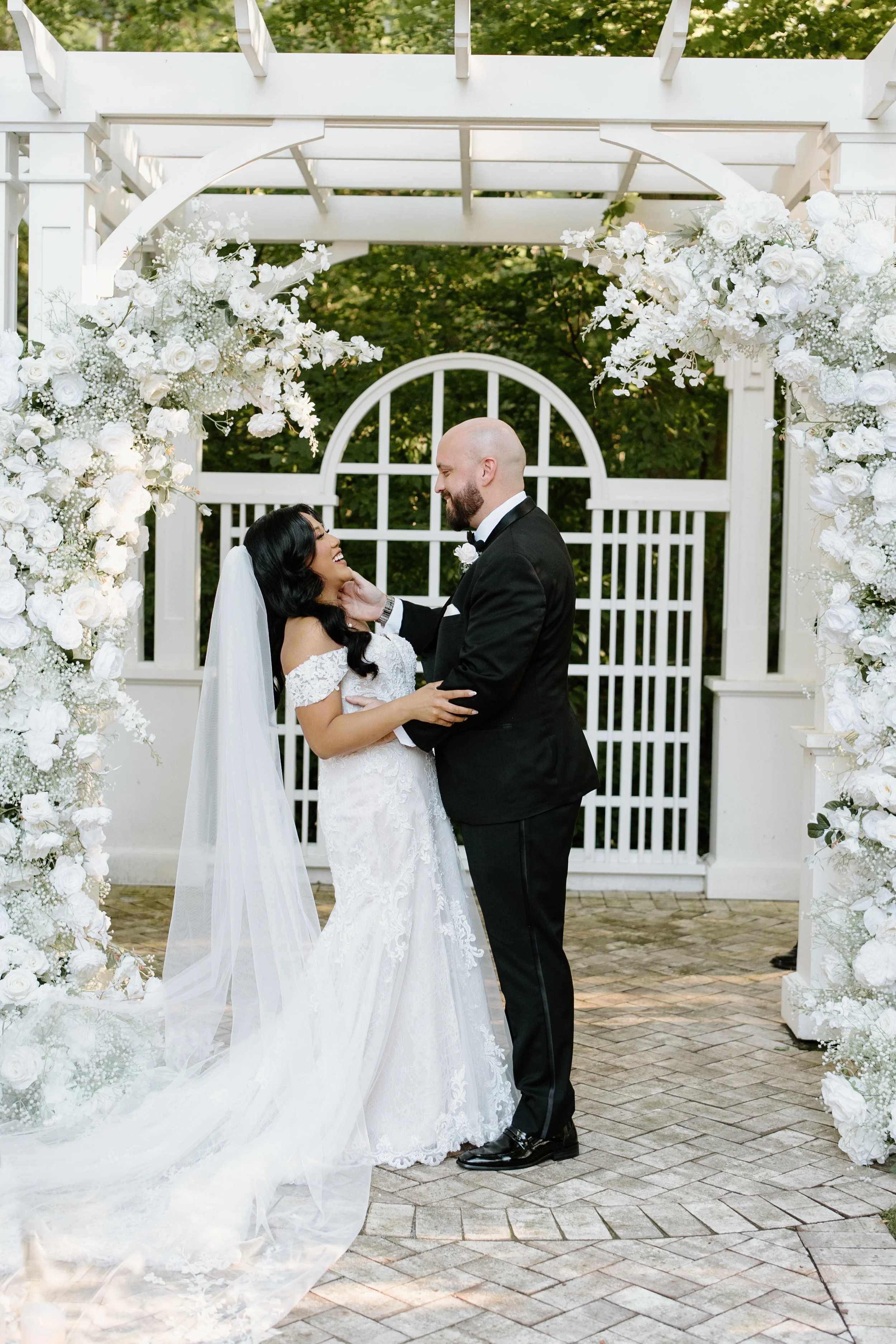 A bride and groom holding hands and gazing at each other under a floral arch at their outdoor wedding ceremony.