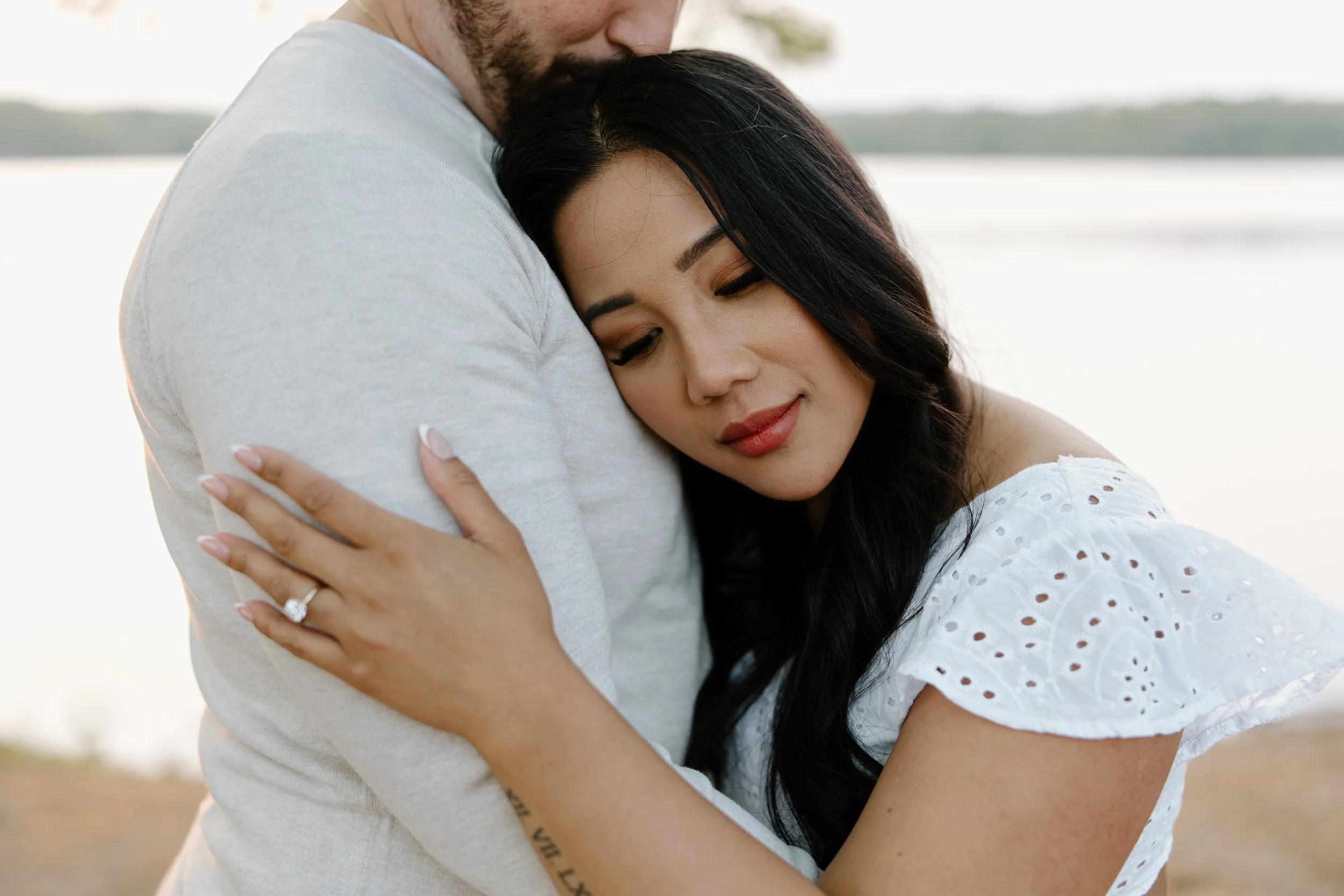 A couple hugging each other near a body of water during sunset, with the woman resting her head on the man's shoulder, both appearing peaceful and content.
