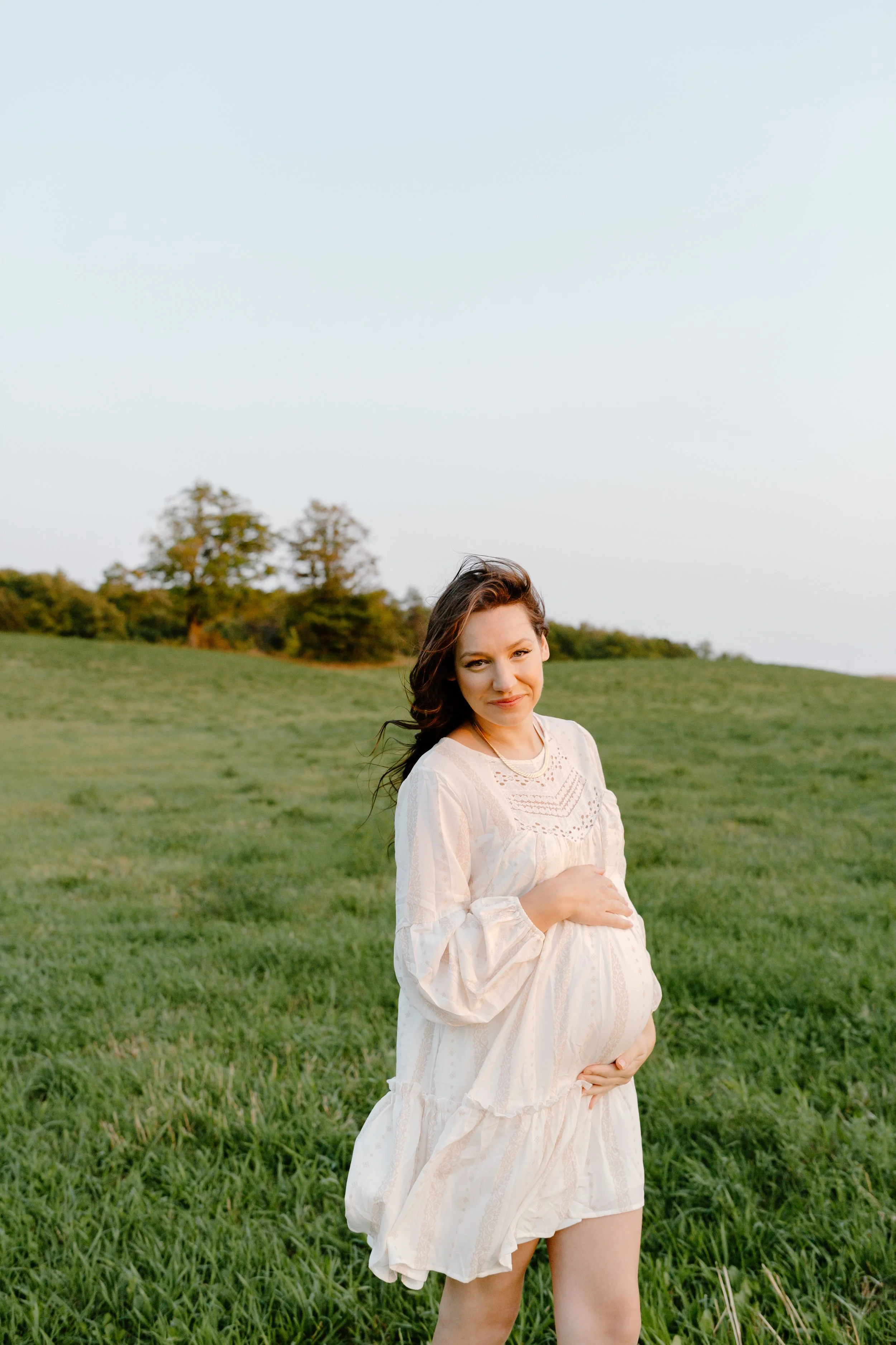 Pregnant woman in a white dress standing in a grassy field during sunset.