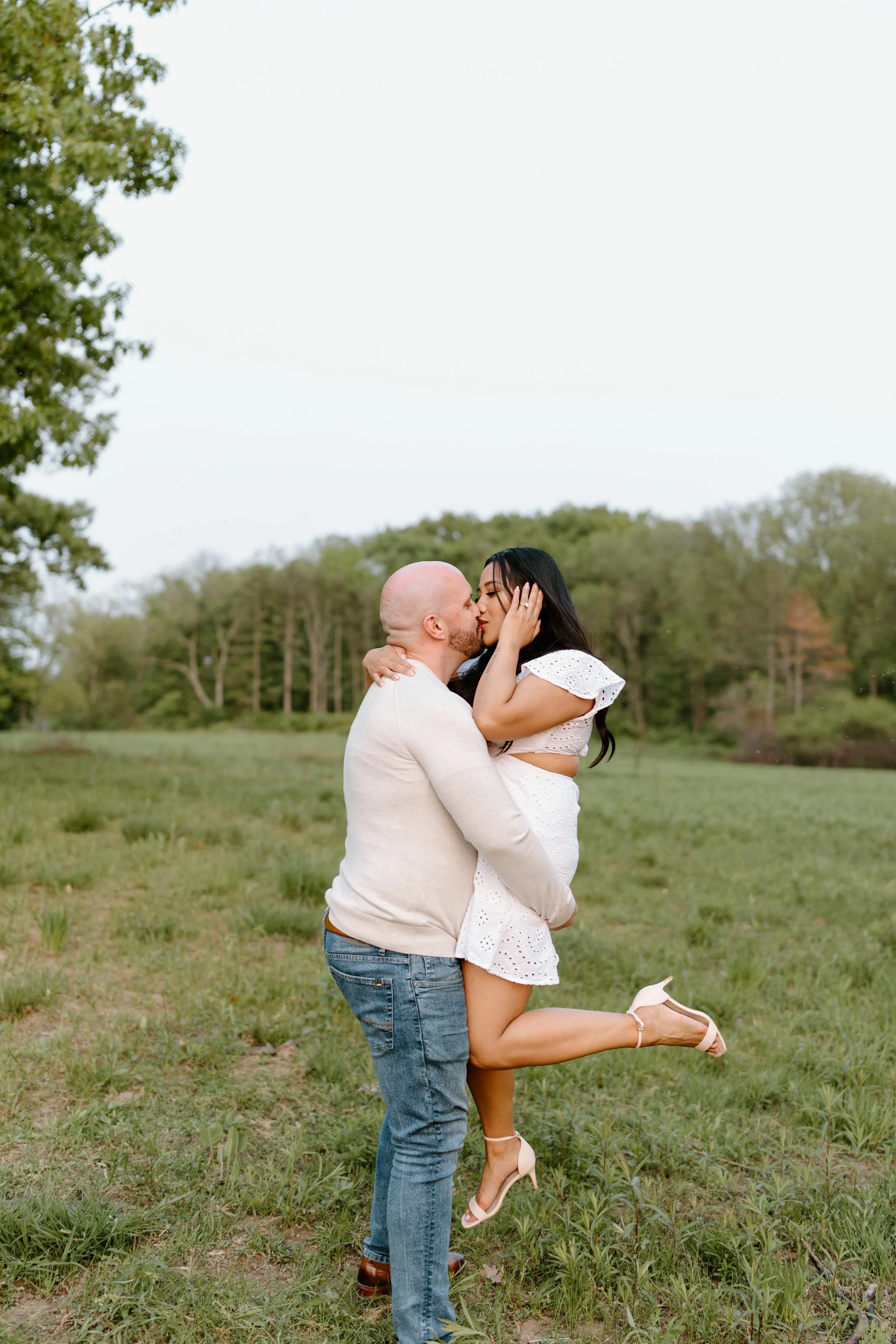 A couple kissing outdoors in a green field with trees in the background. The man is holding the woman, who is wearing a white dress and high heels, while the woman has her eyes closed and one leg bent.