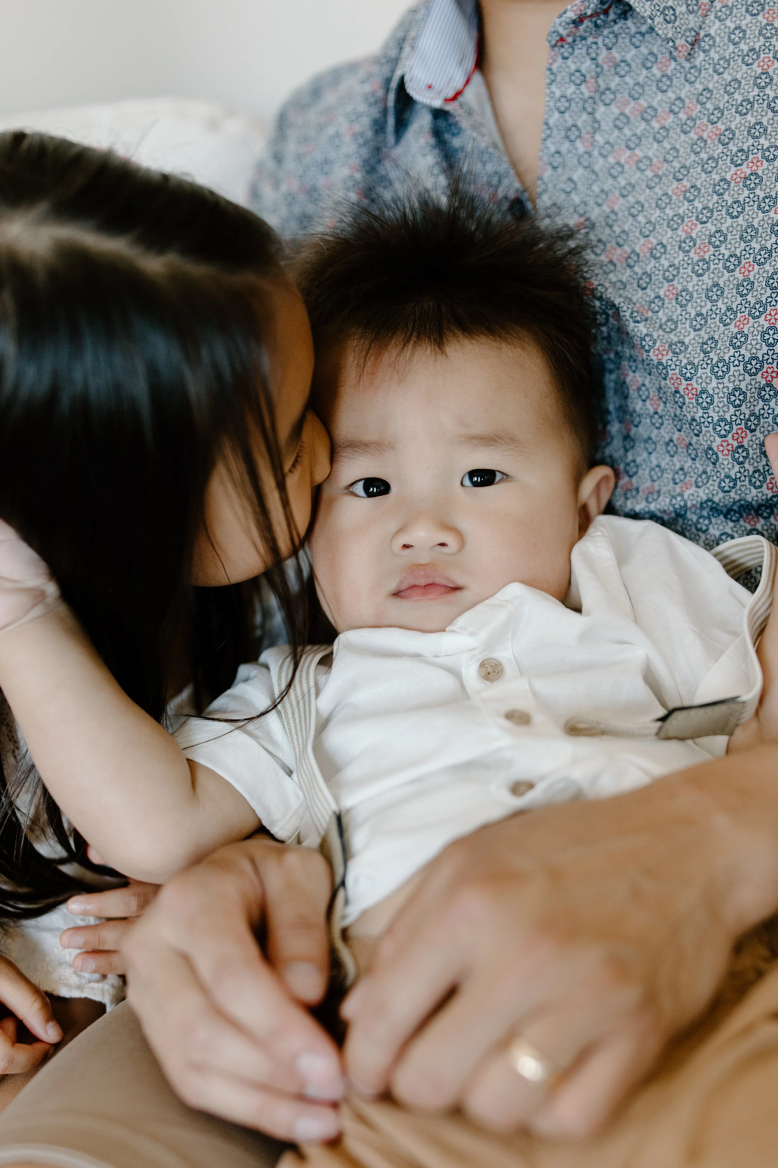 A young girl is kissing a young boy on the face while sitting close to him on a couch. The boy appears to be looking at the camera with a neutral expression, and an adult's hand holds his hand.
