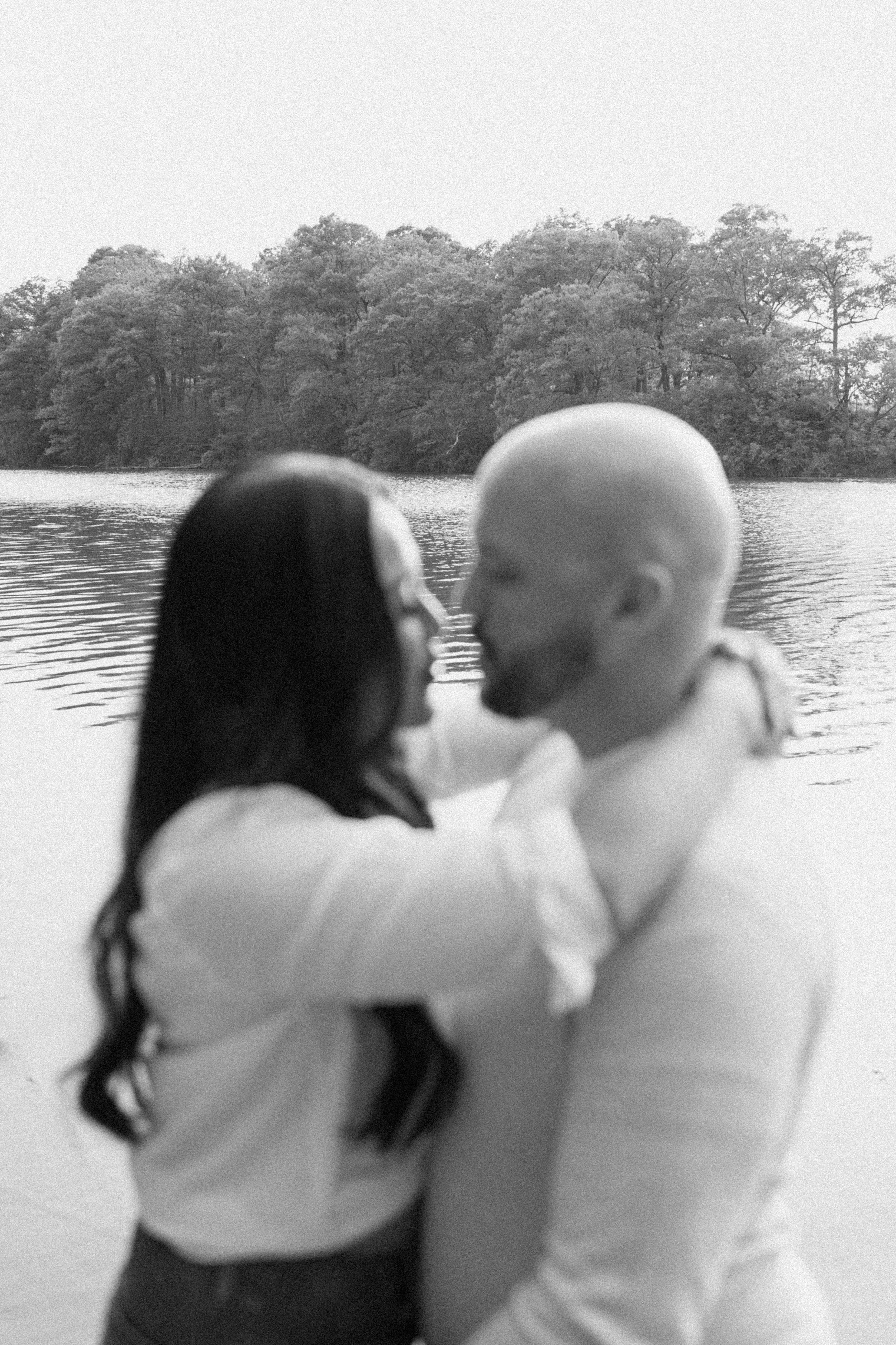A couple embraces near a body of water with trees in the background.