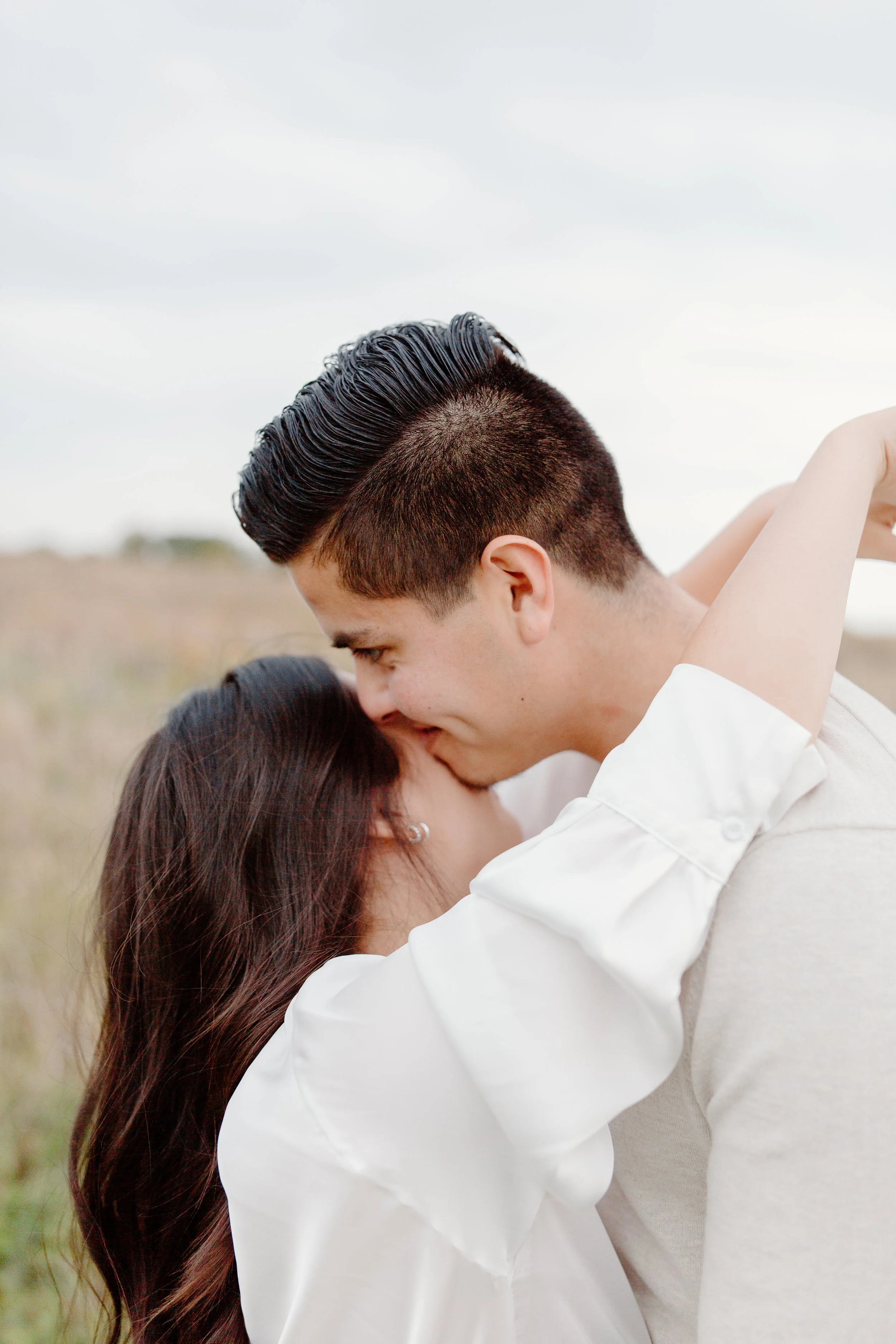 A couple sharing a kiss outdoors, with the man holding the woman close, both smiling, in a natural setting with a cloudy sky.