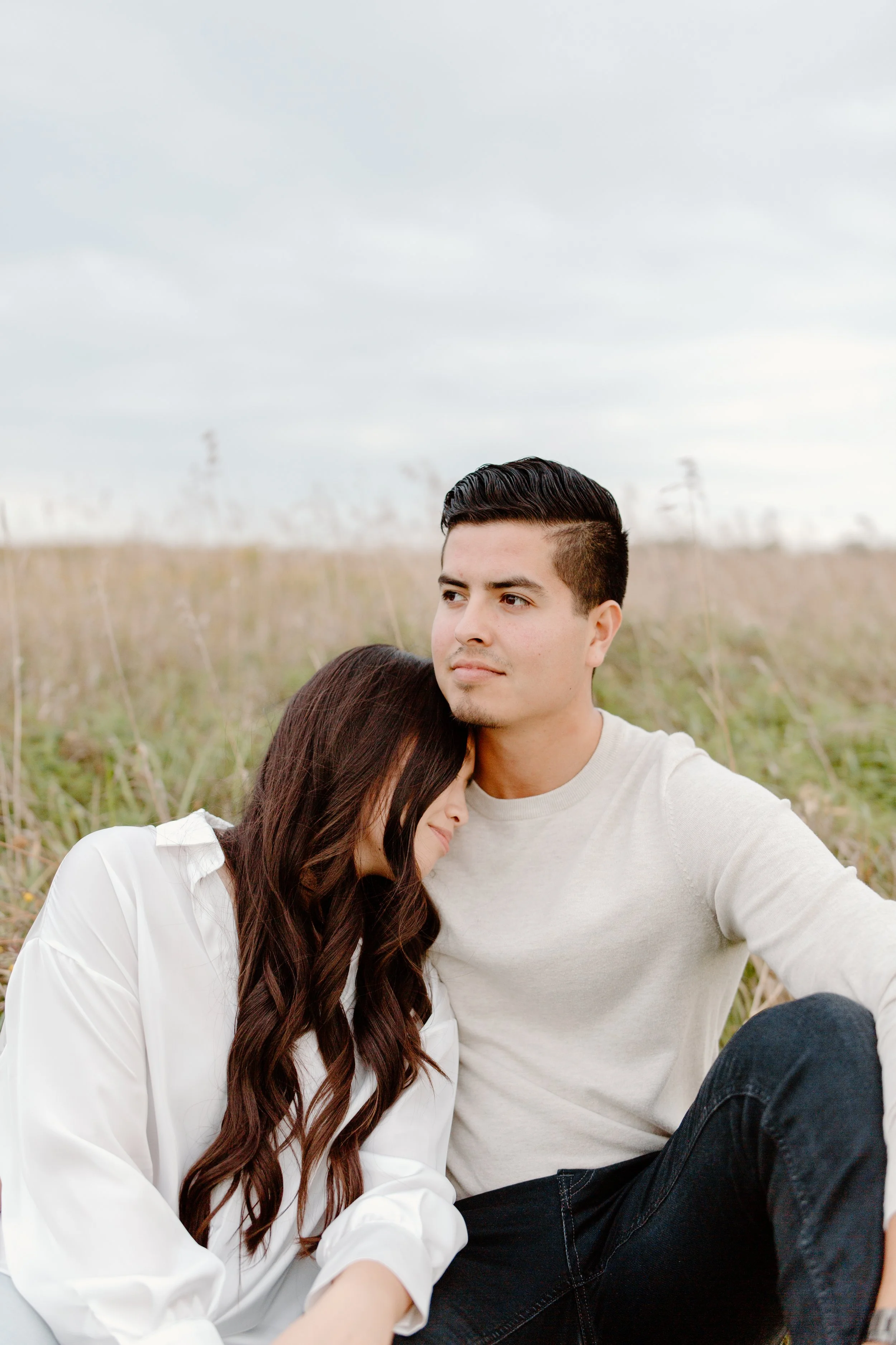 A young couple sitting in a field, with the woman resting her head on the man's shoulder, wearing white and grey clothes.
