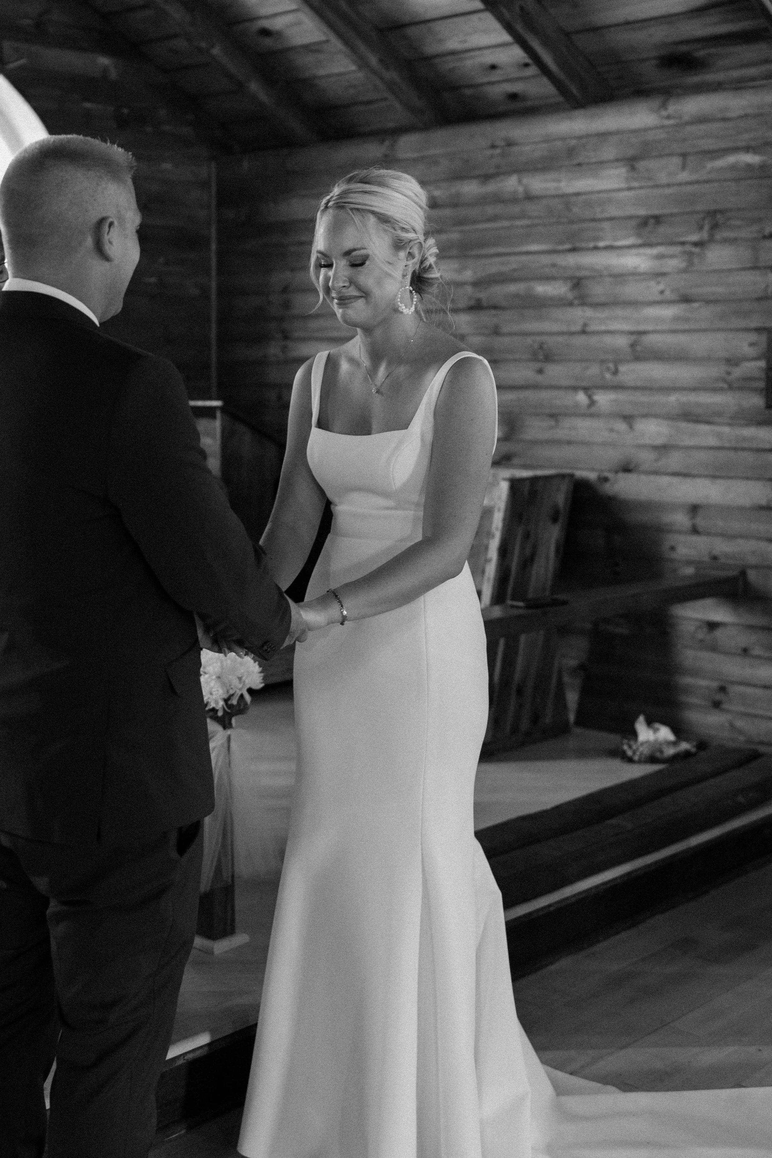 A woman in a white wedding gown and earrings is holding hands with a man in a suit during a wedding ceremony inside a wooden venue.