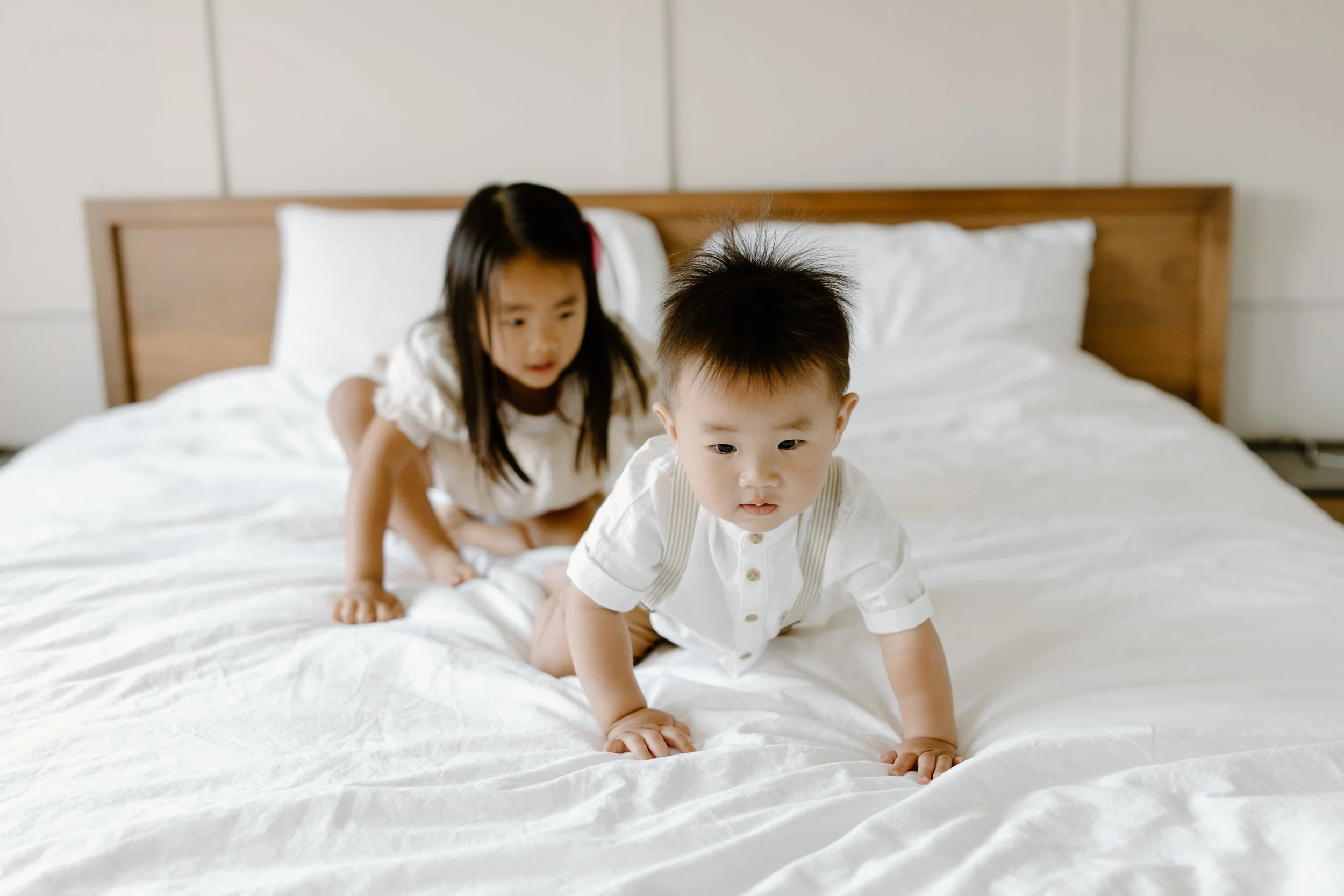 Two young children, a girl and a boy, crawling on a white bed with a wooden headboard in a bright room.