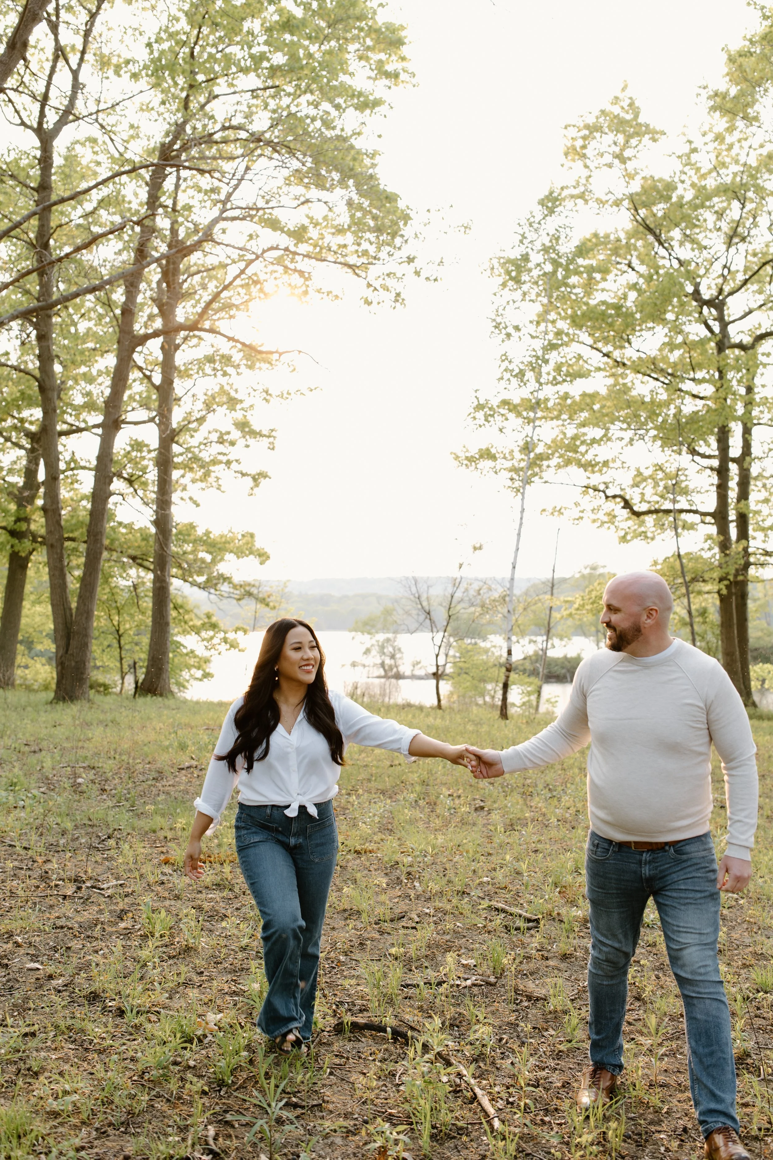 A couple holding hands and walking in a wooded area near a lake during sunset.