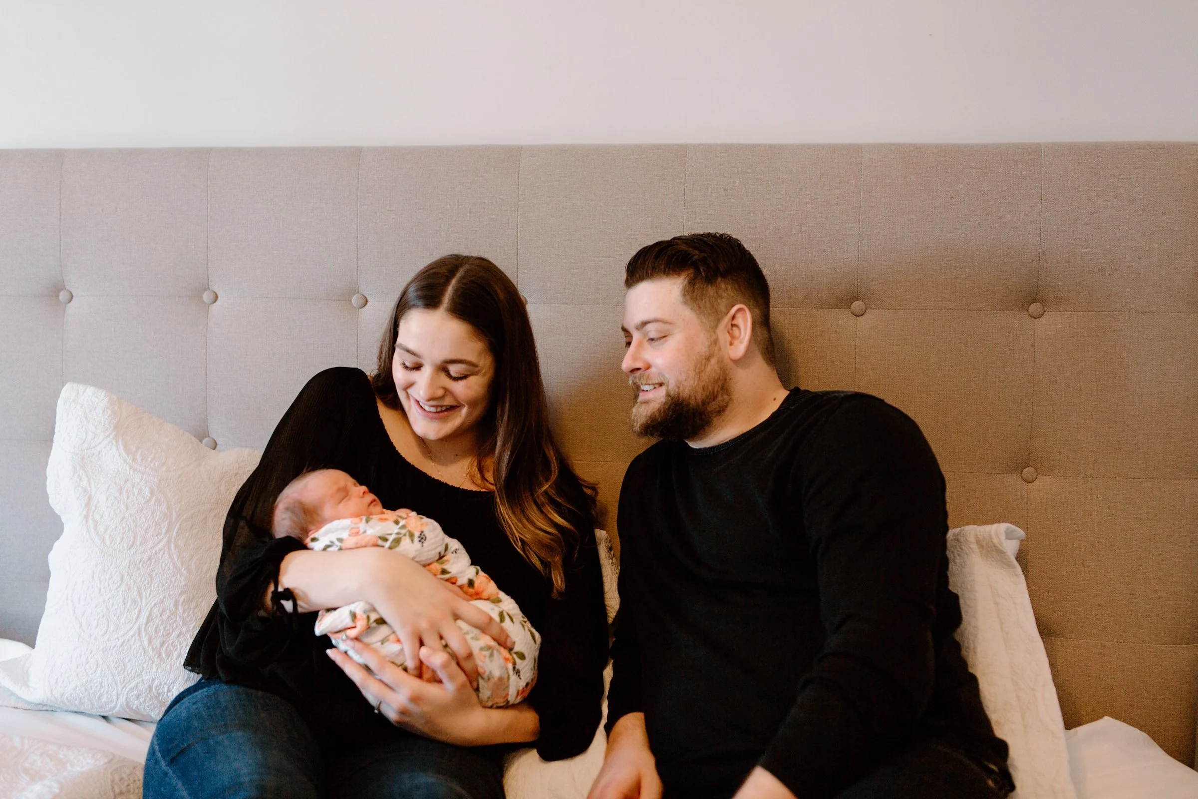 A smiling woman holding a newborn baby, sitting on a bed with a man next to her, in a warmly lit bedroom.