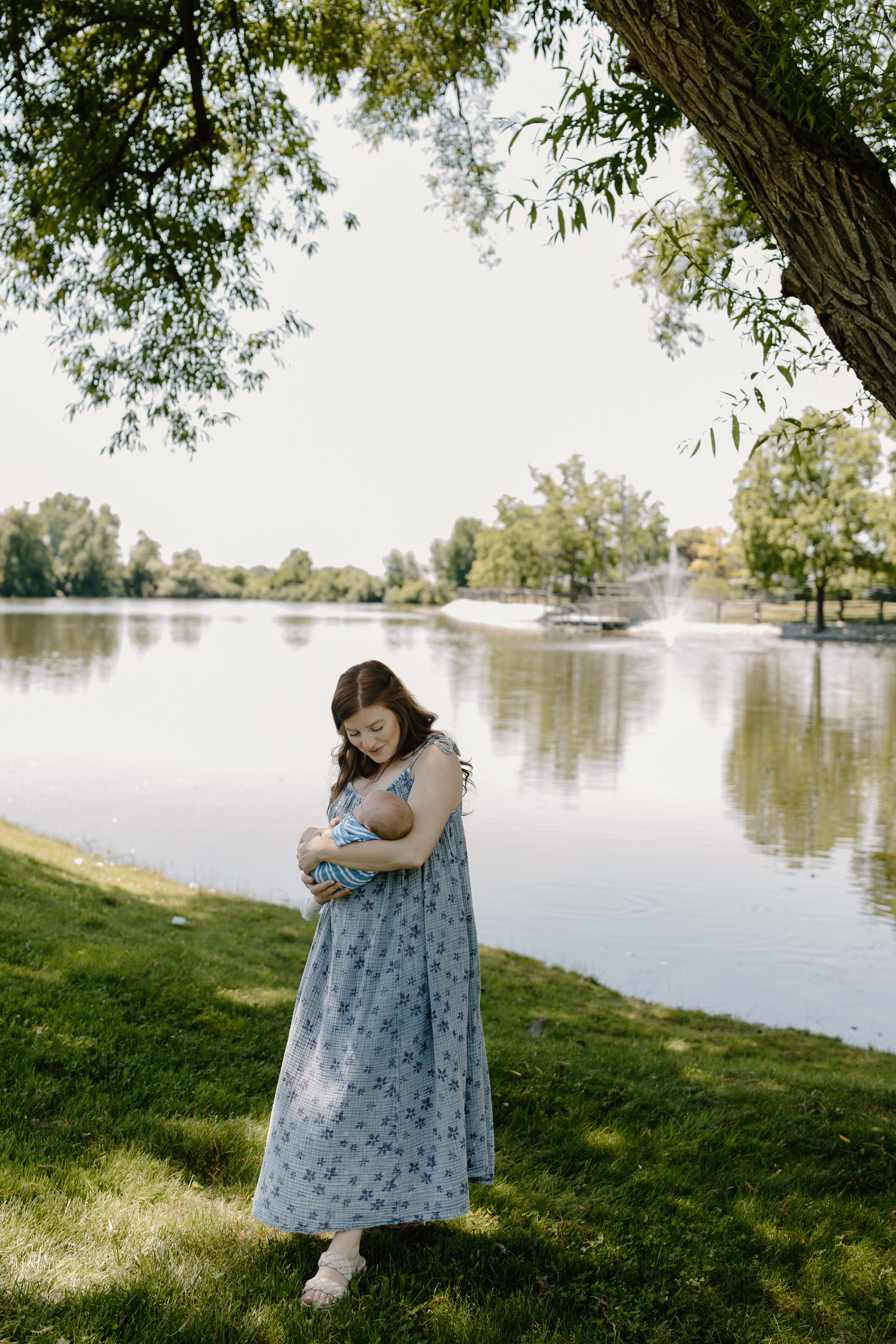 A woman in a long floral dress holding a baby near a lake under a large tree, with a fountain in the background.