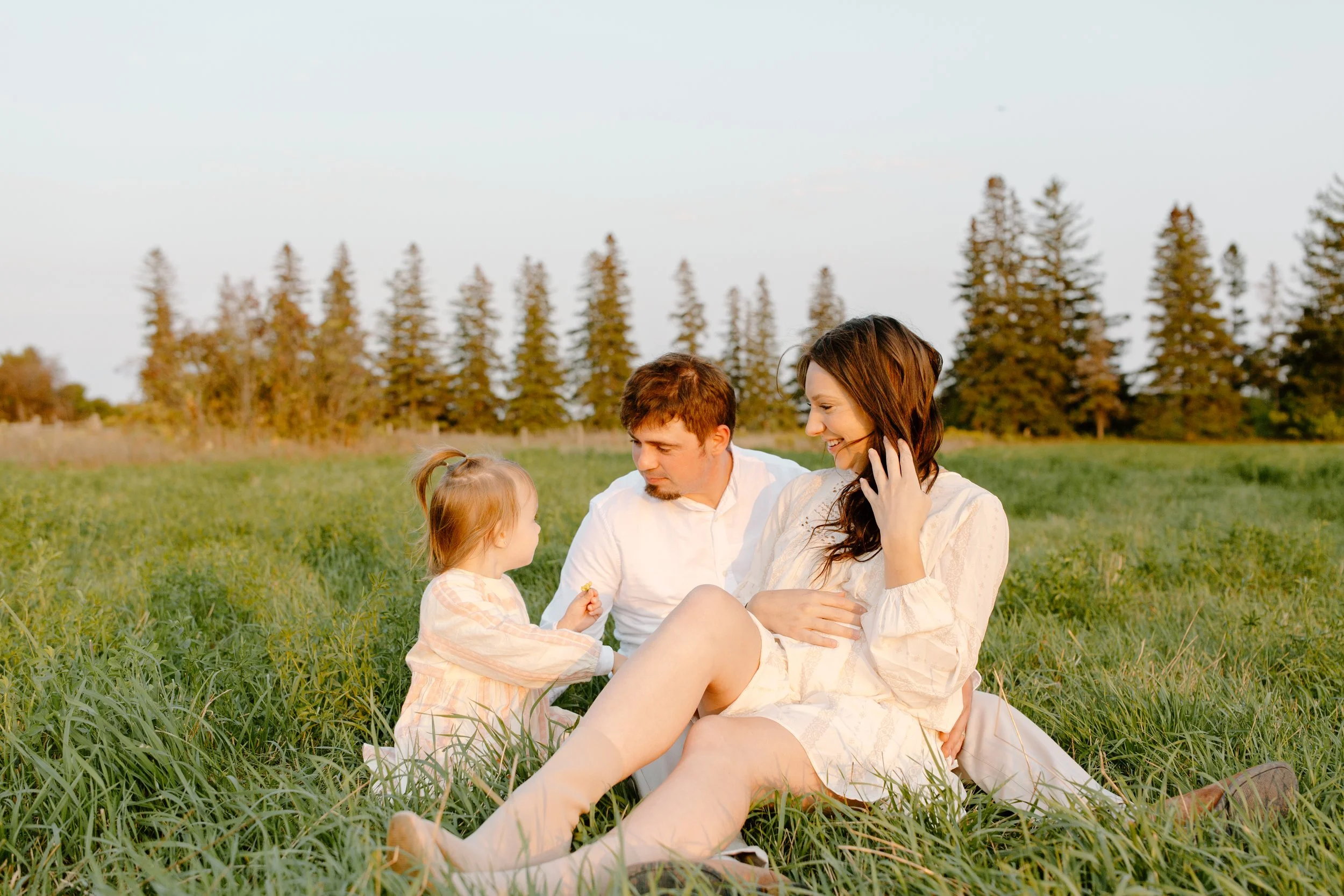A family of three sitting on grass in a field, with trees in the background during sunset. The mother, father, and young daughter are interacting happily, with the mother smiling and the father looking at the daughter.