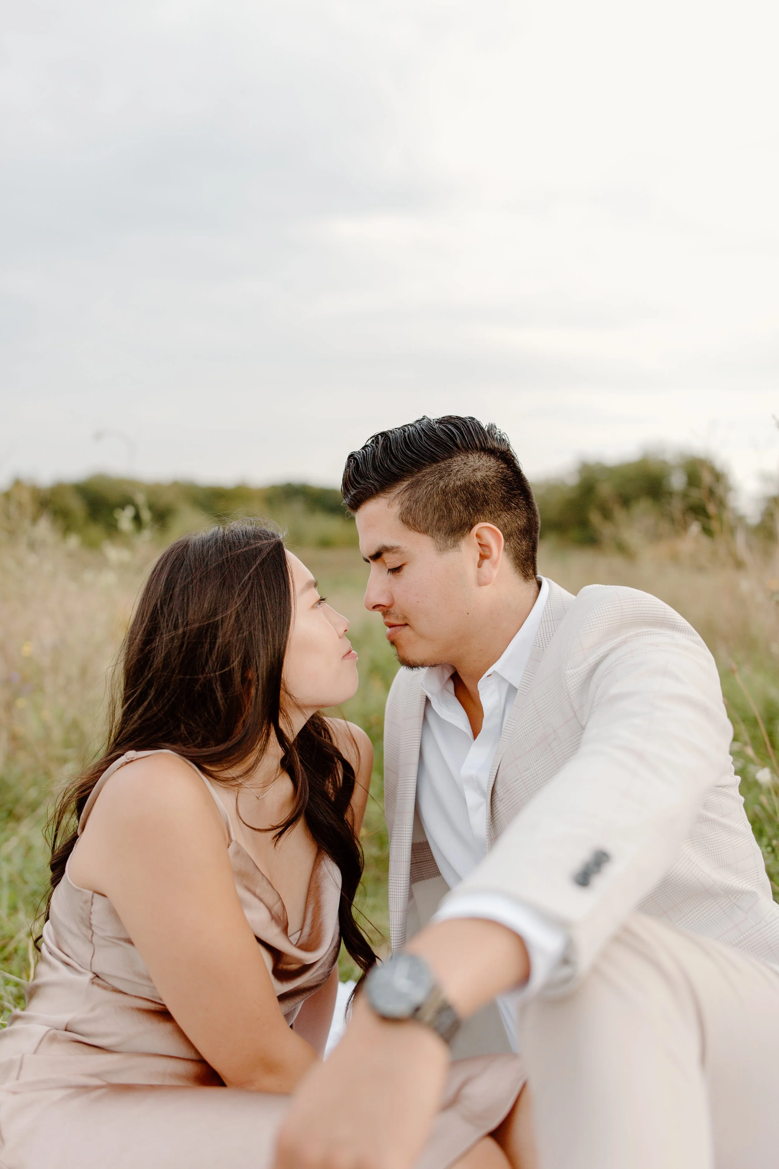 A couple sitting close together outdoors in a field, with noses nearly touching, and a cloudy sky overhead.