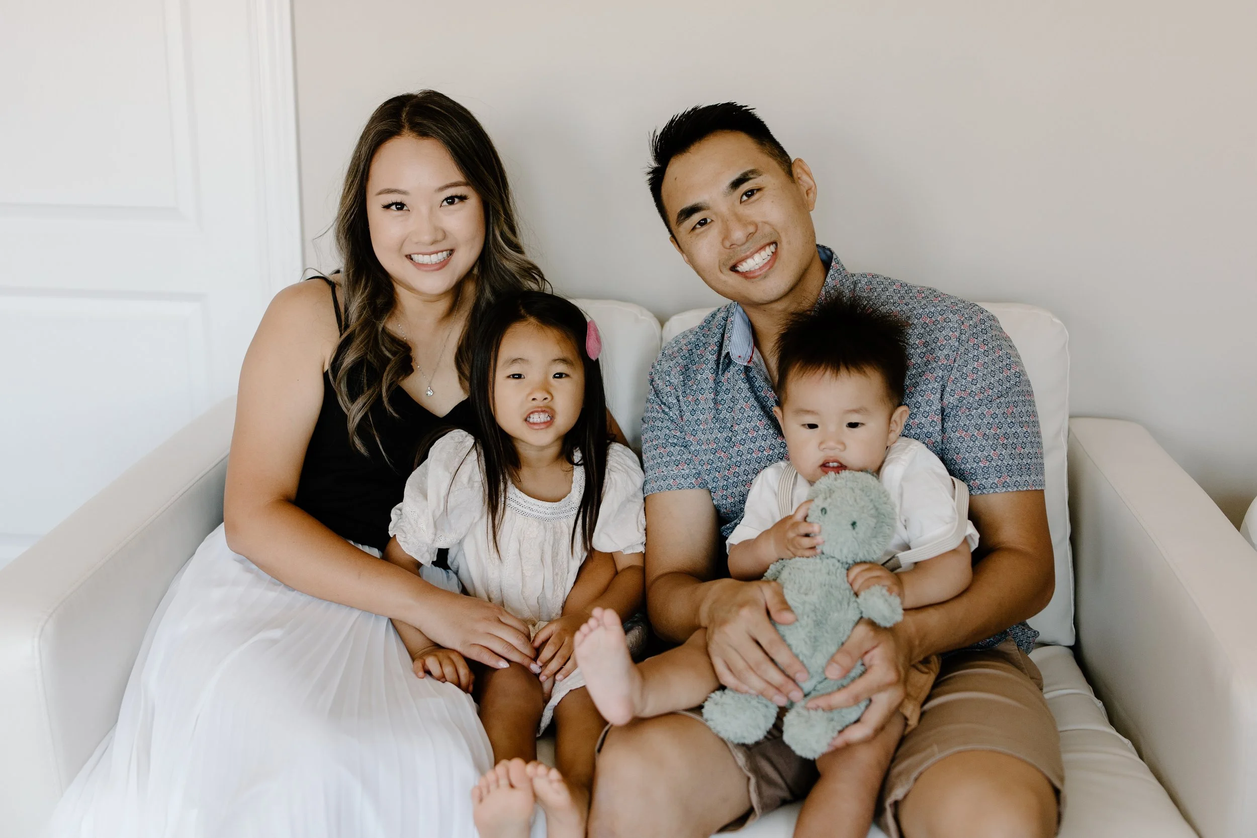 A happy family of four sitting on a white sofa in a bright room. The mother, with long wavy hair, wearing a black top and white skirt, is smiling. The father, with short black hair, wearing a gray patterned shirt, is smiling. The young girl, with lon