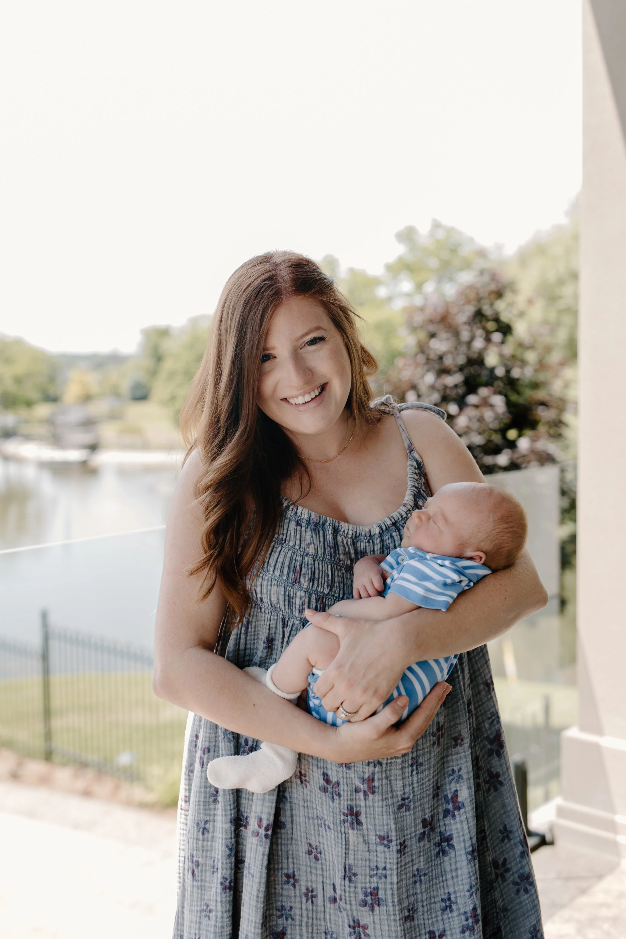A woman with long brown hair smiling and holding a sleeping baby dressed in a blue and white striped outfit outdoors near a body of water and greenery.