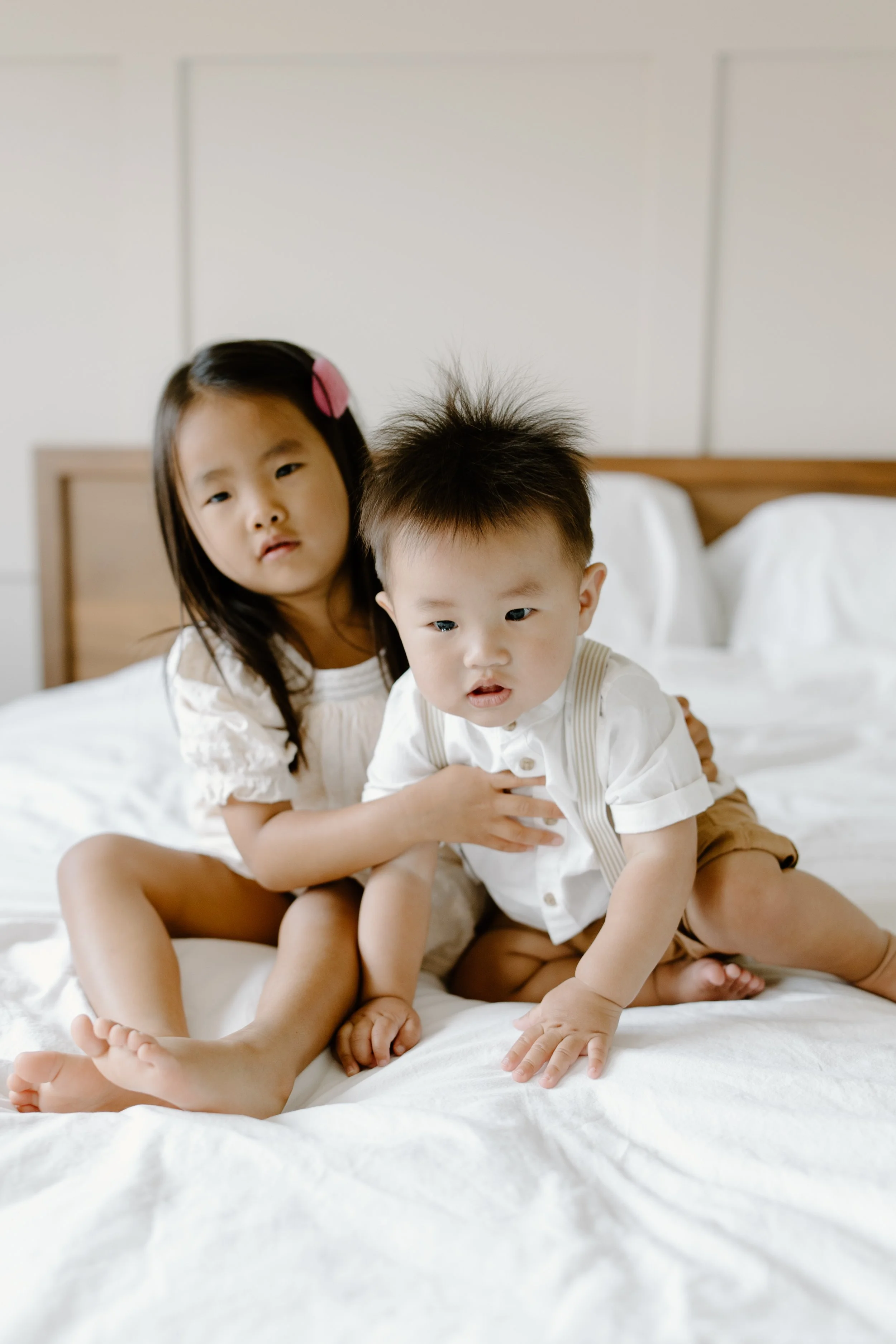 Two young children, a girl and a boy, sitting on a bed in a bedroom. The girl has long dark hair with a pink clip and is wearing a white dress. The boy has spiky hair, wearing a white shirt and tan shorts.