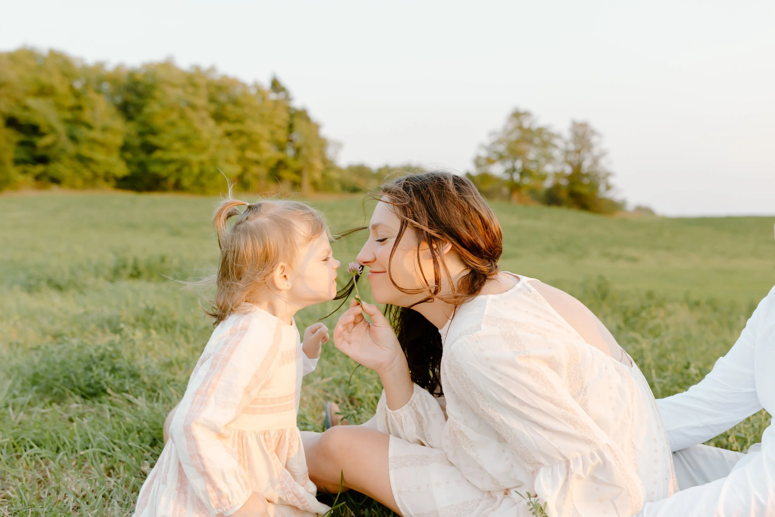 A woman and a young girl sitting on grass outdoors, touching noses, with the girl holding a flower near the woman's face, in a green field with trees in the background.