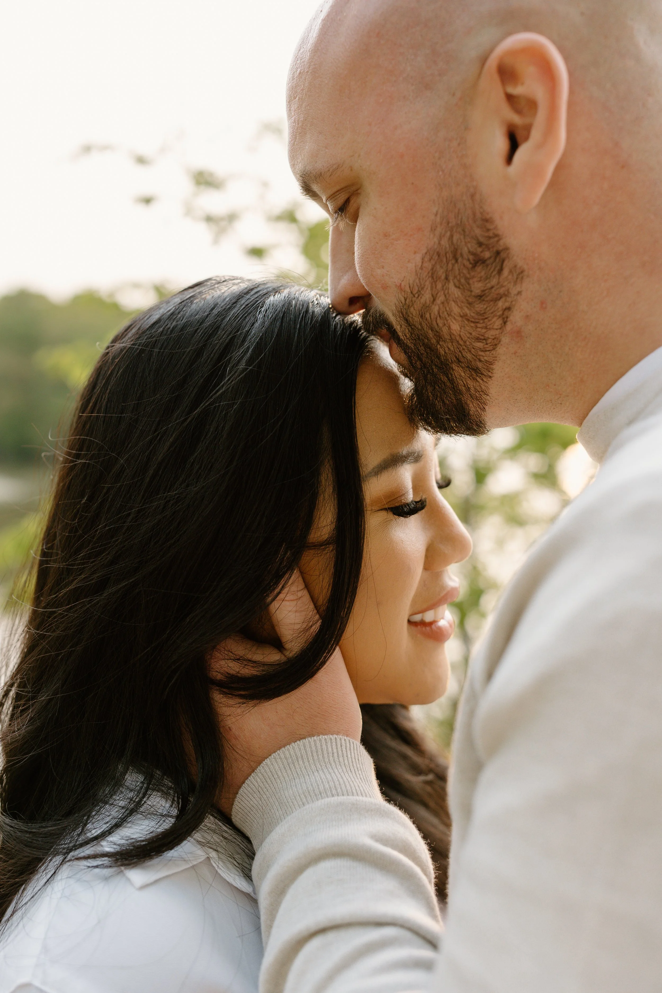 A close-up image of a man and woman with eyes closed, sharing an intimate moment outdoors. The man is gently kissing the woman's forehead while holding her face, and the woman is smiling with her head slightly tilted back.