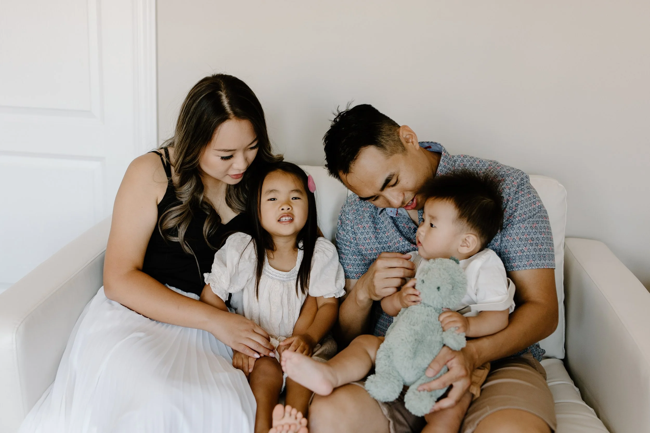 A family of five sitting on a white couch in a living room, with two young children, a girl and a boy, and their parents, engaging in a close and tender moment.