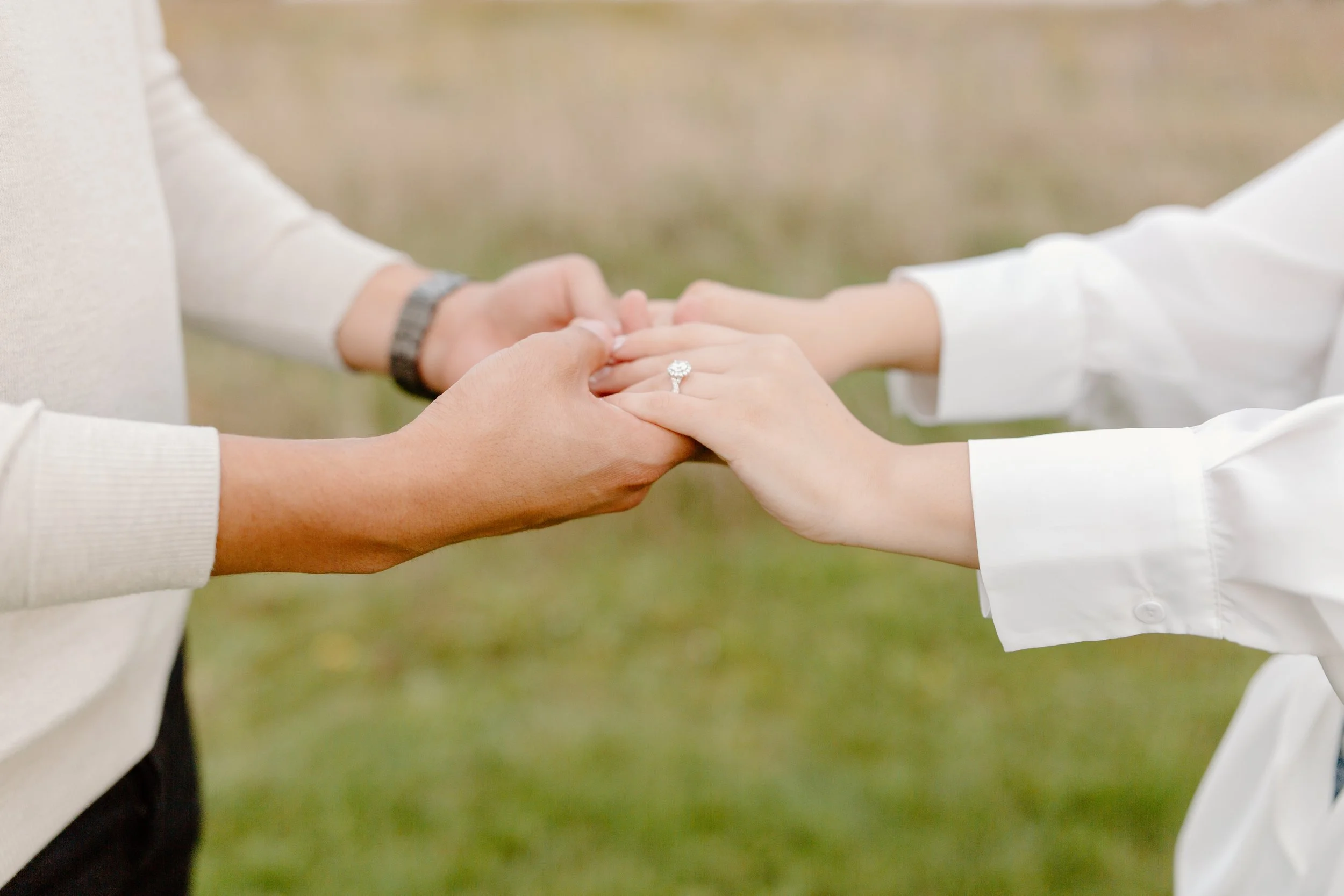 Two people are holding hands, with a ring visible on the finger of the person on the right. They are outdoors, on a grassy area, and appear to be embracing or exchanging a gesture of affection.