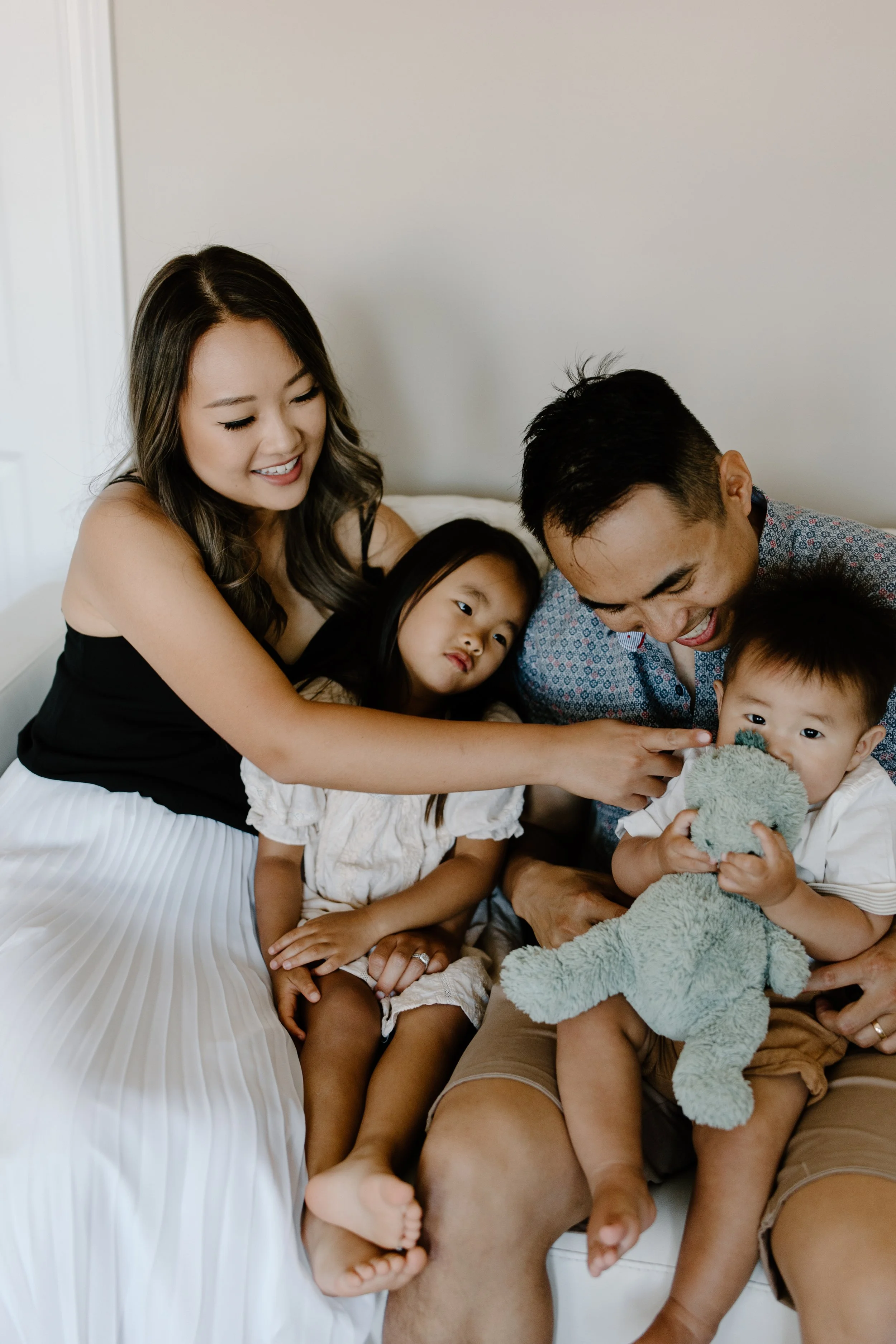 A family of four, including two young children, sits together on a white sofa. The children are holding plush toys, and the parents are smiling and engaging with the kids, creating a warm, joyful atmosphere.