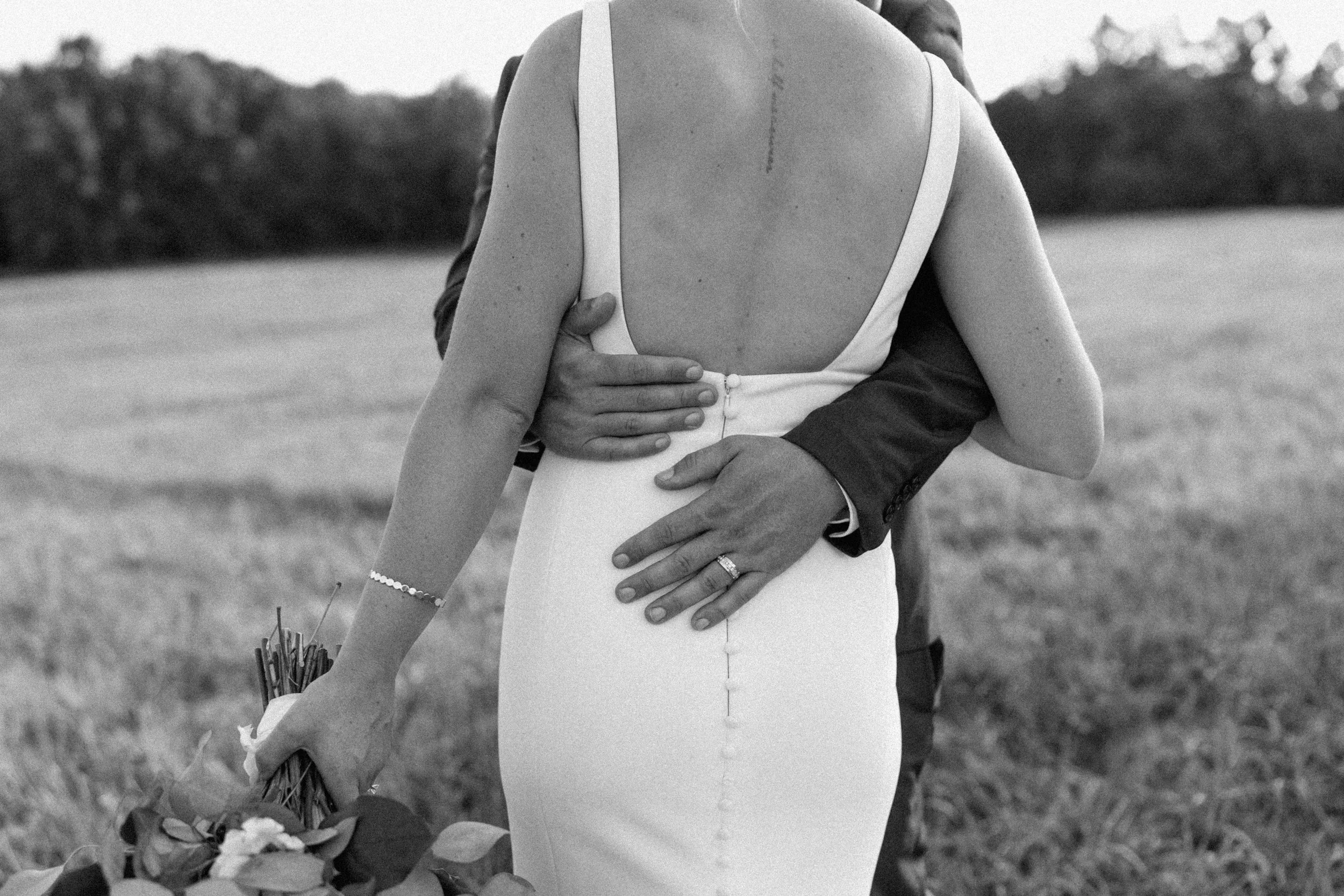 A couple at their wedding, with the woman's back to the camera and a man embracing her from behind, outdoors in a field.