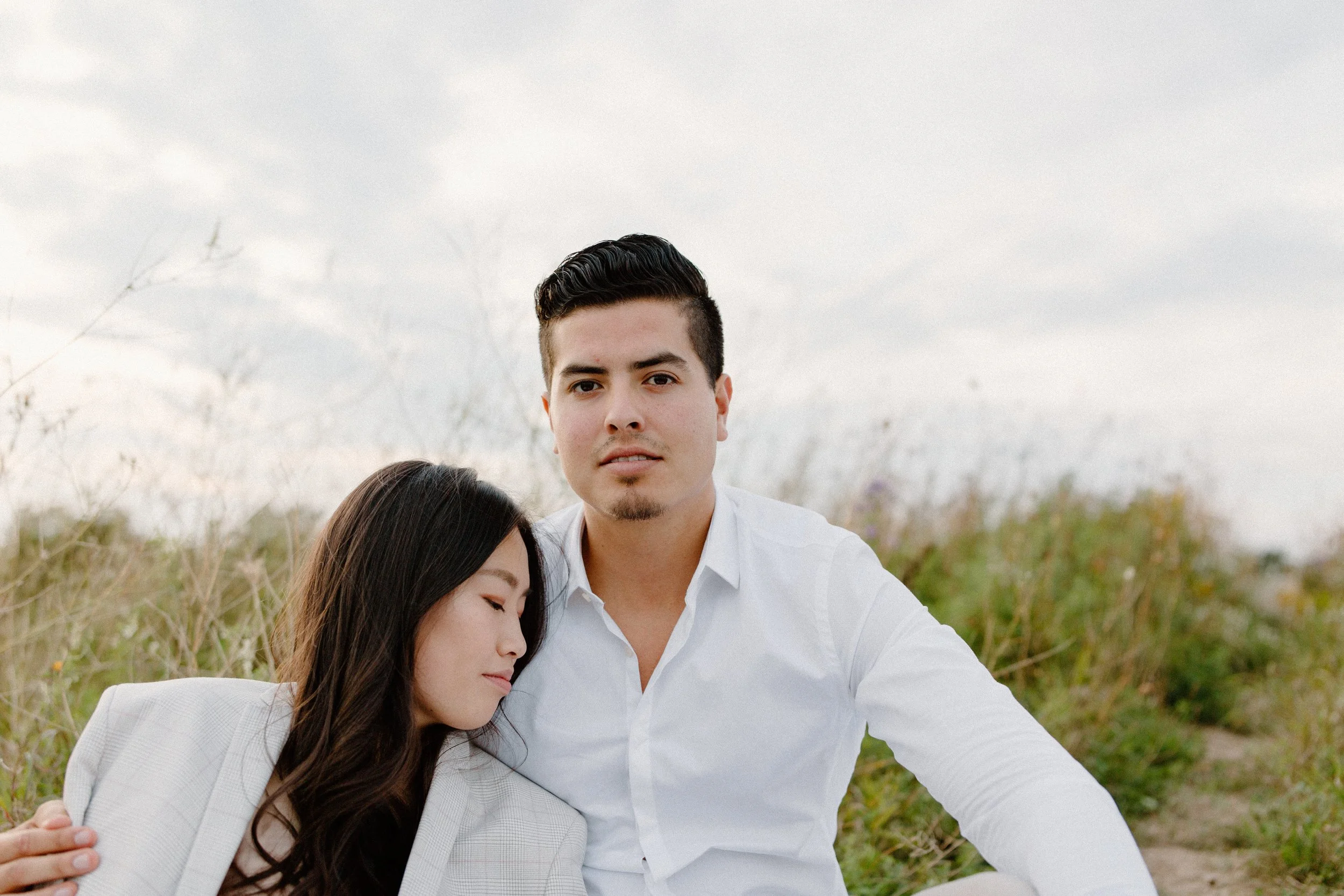 A man and woman sit outdoors in a field during daytime, with the woman resting her head on the man's shoulder. The man wears a white shirt and looks directly at the camera, while the woman has long dark hair and eyes closed.