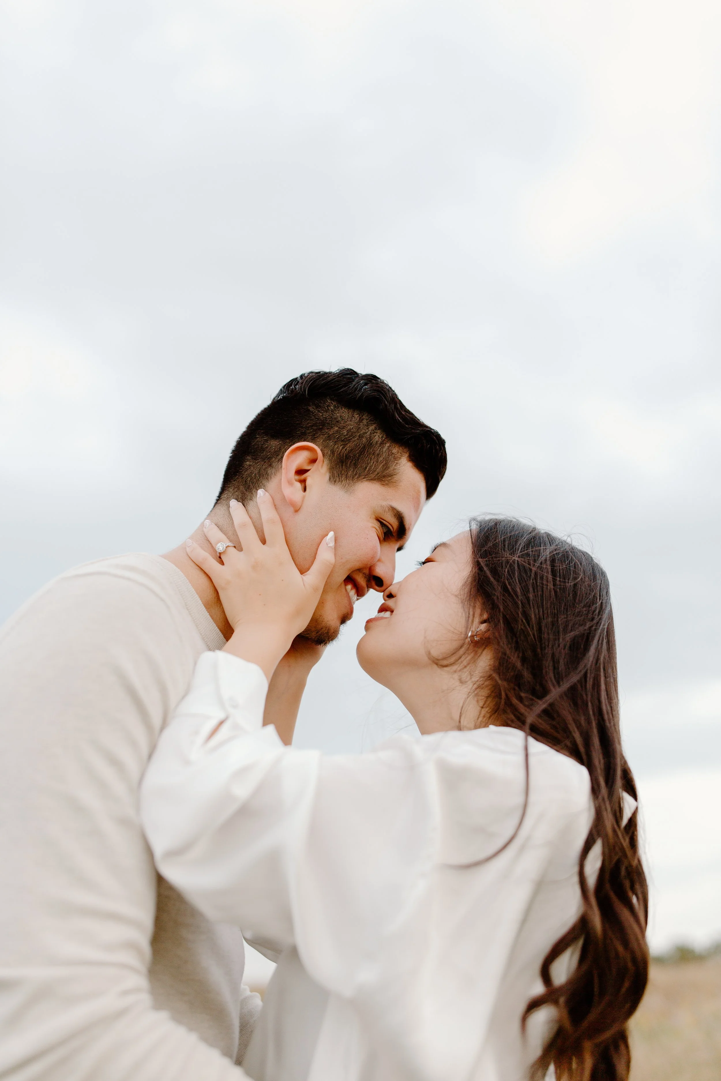 A couple is touching foreheads, smiling, outdoors with a cloudy sky behind them.