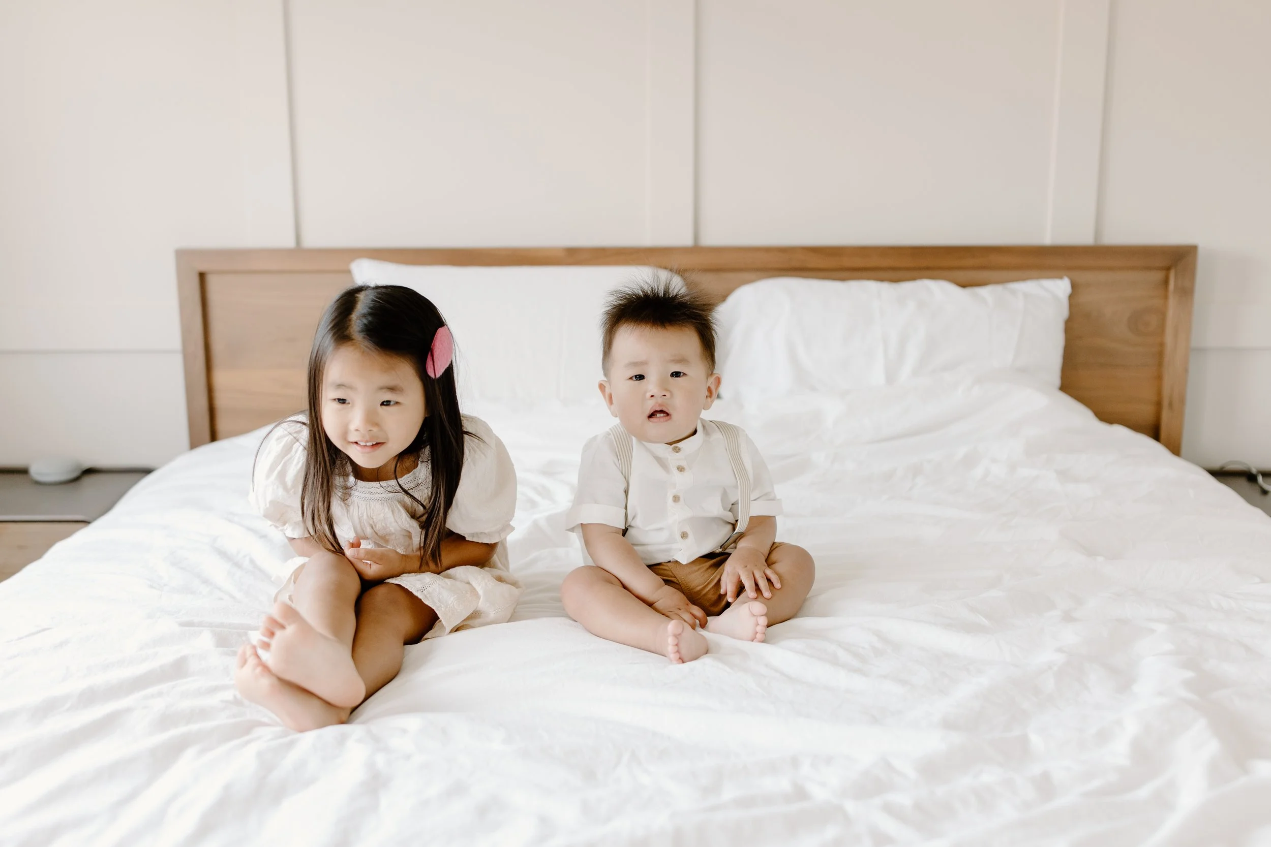 Two young children sitting on a white bed in a minimalist bedroom.