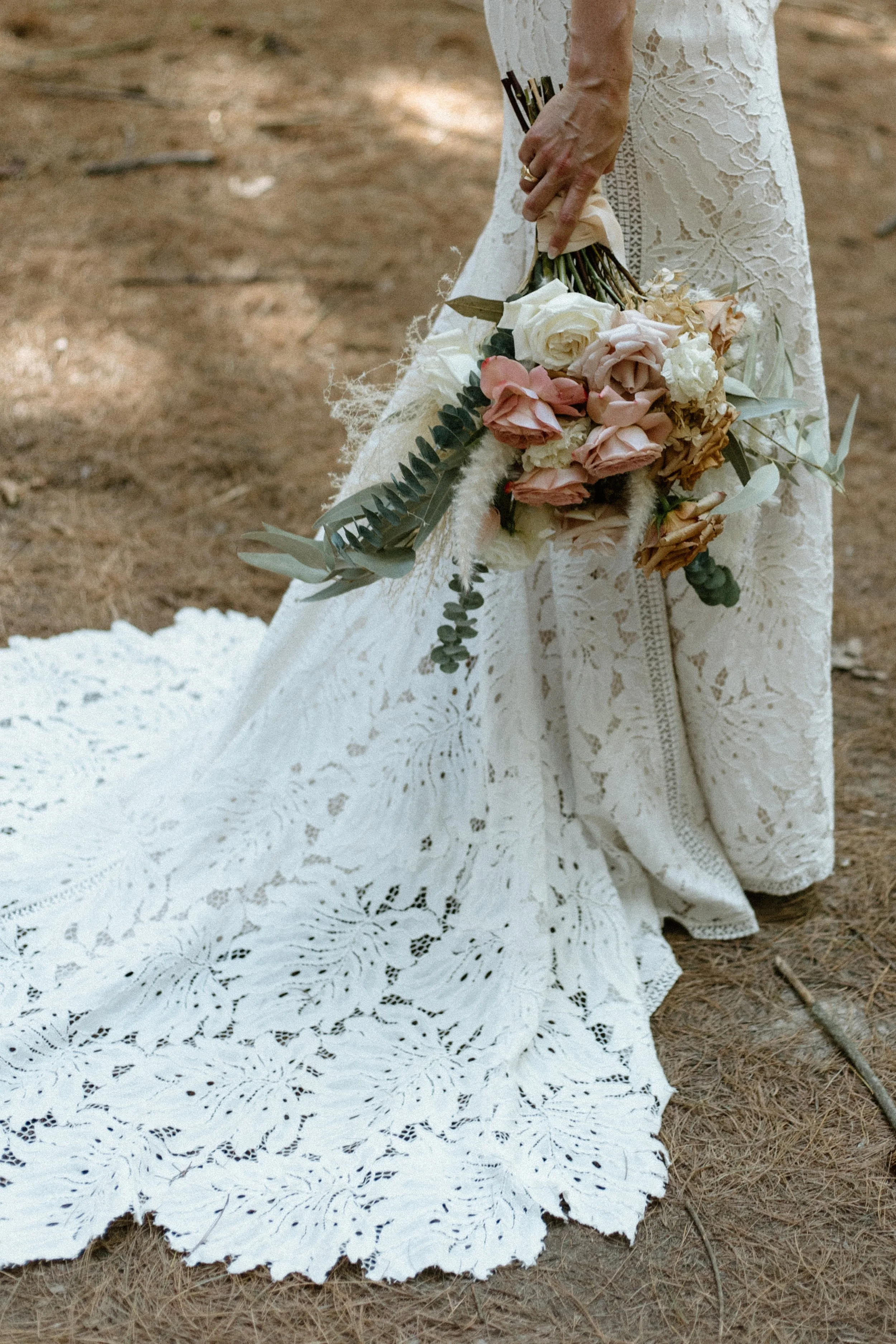 A bride holding a bouquet of pink, cream, and peach flowers, standing on a lace train on the ground.