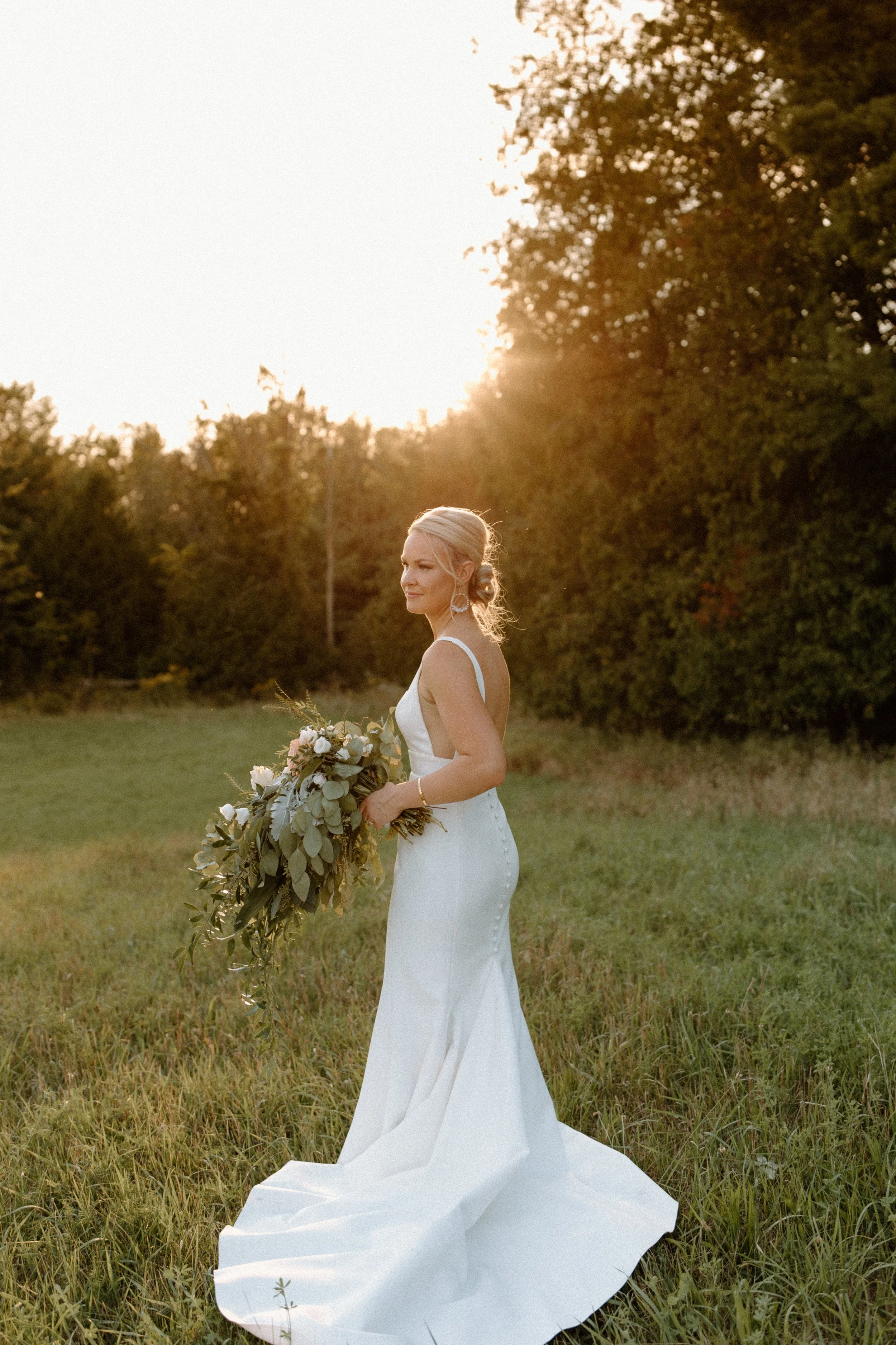 A bride in a white wedding dress holding a bouquet of flowers outdoors during sunset.