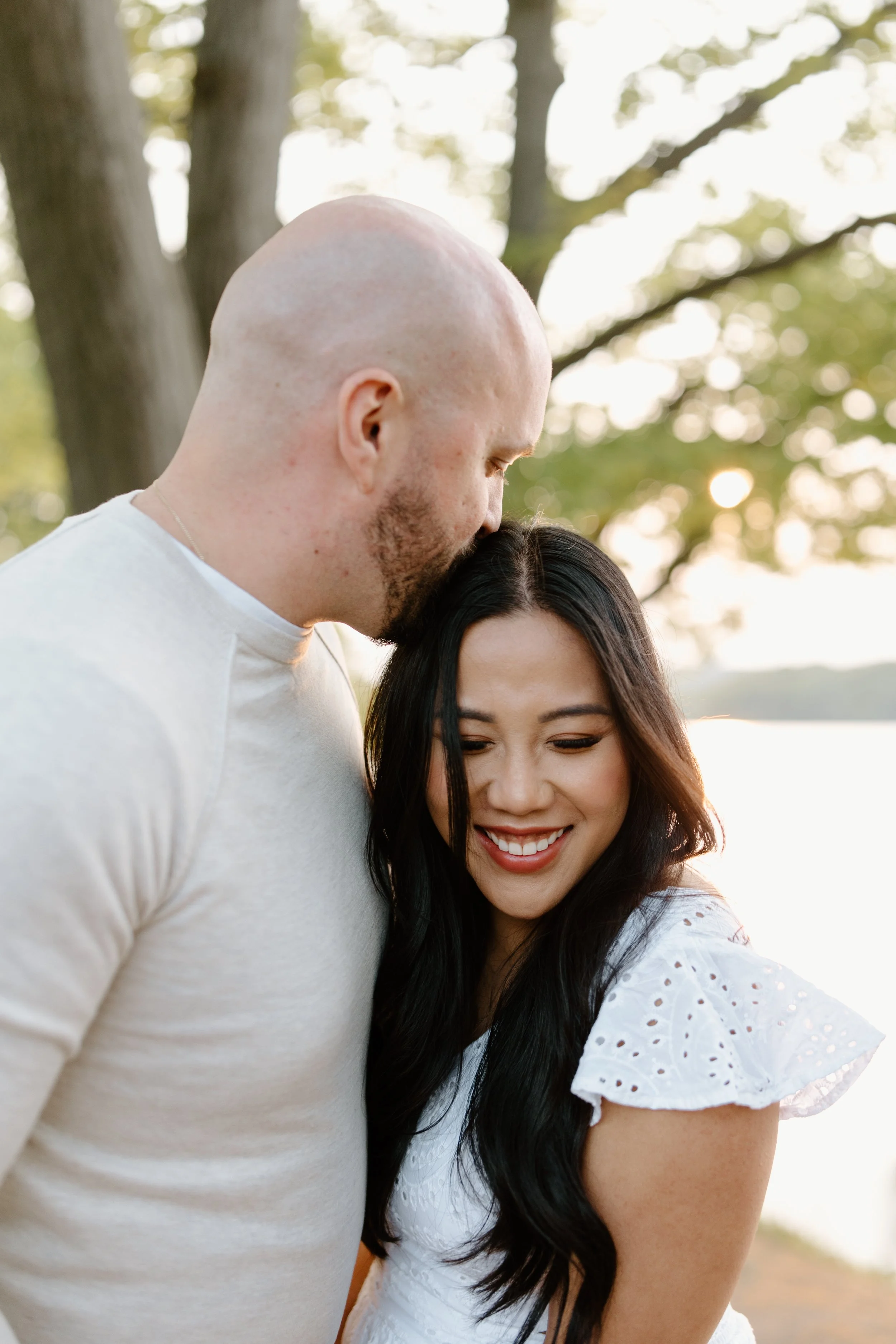 A couple standing outdoors by a lake, with trees in the background, sharing a tender moment as the man kisses the woman on her head.