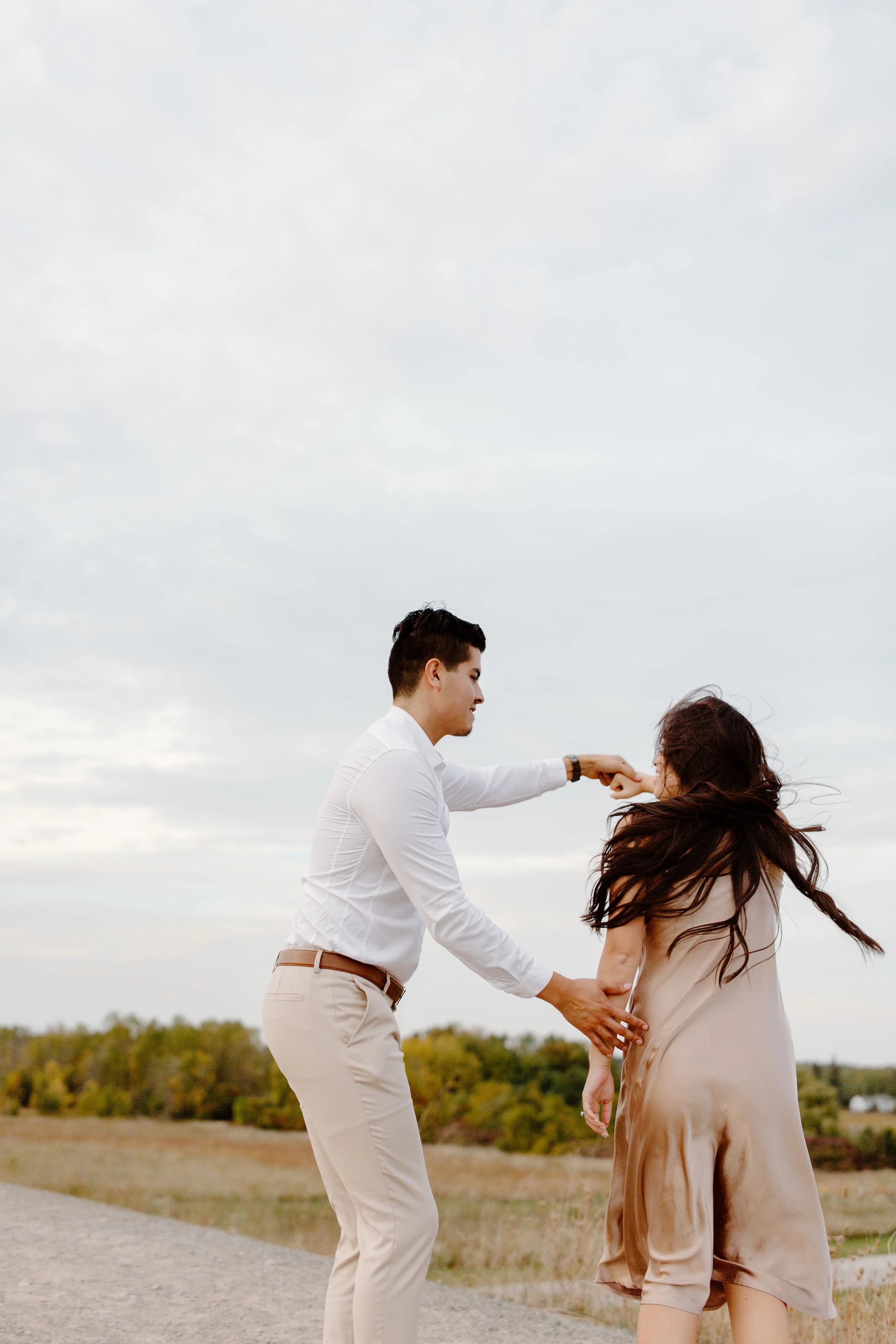 A couple is dancing outdoors on a cloudy day, with the man holding the woman's hand and the woman’s hair flowing in the wind.