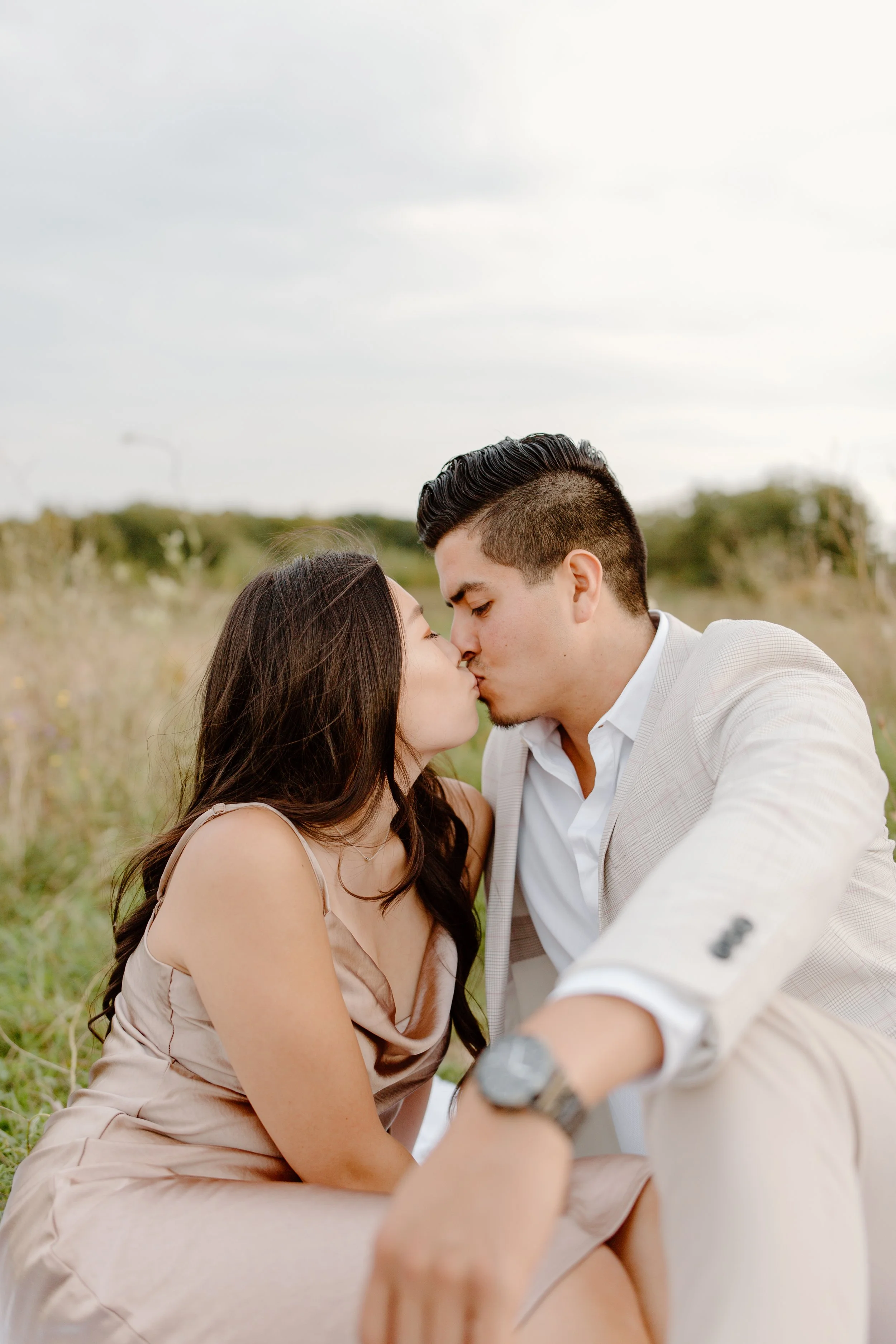 A couple kissing outdoors in a grassy field, dressed in semi-formal attire, with the woman wearing a beige dress and the man in a light-colored suit jacket.