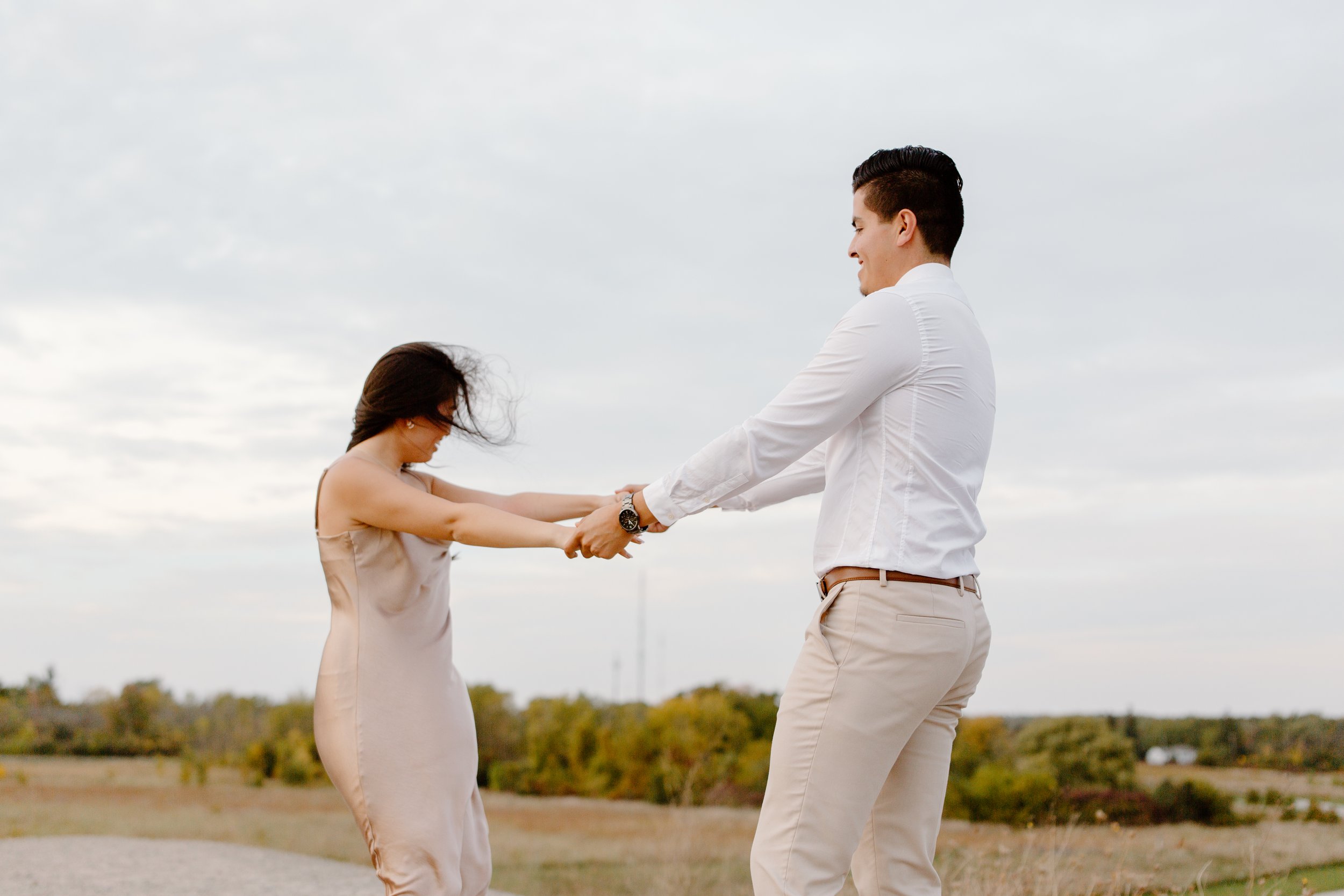A couple standing outdoors holding hands and dancing, with a cloudy sky and fields in the background.