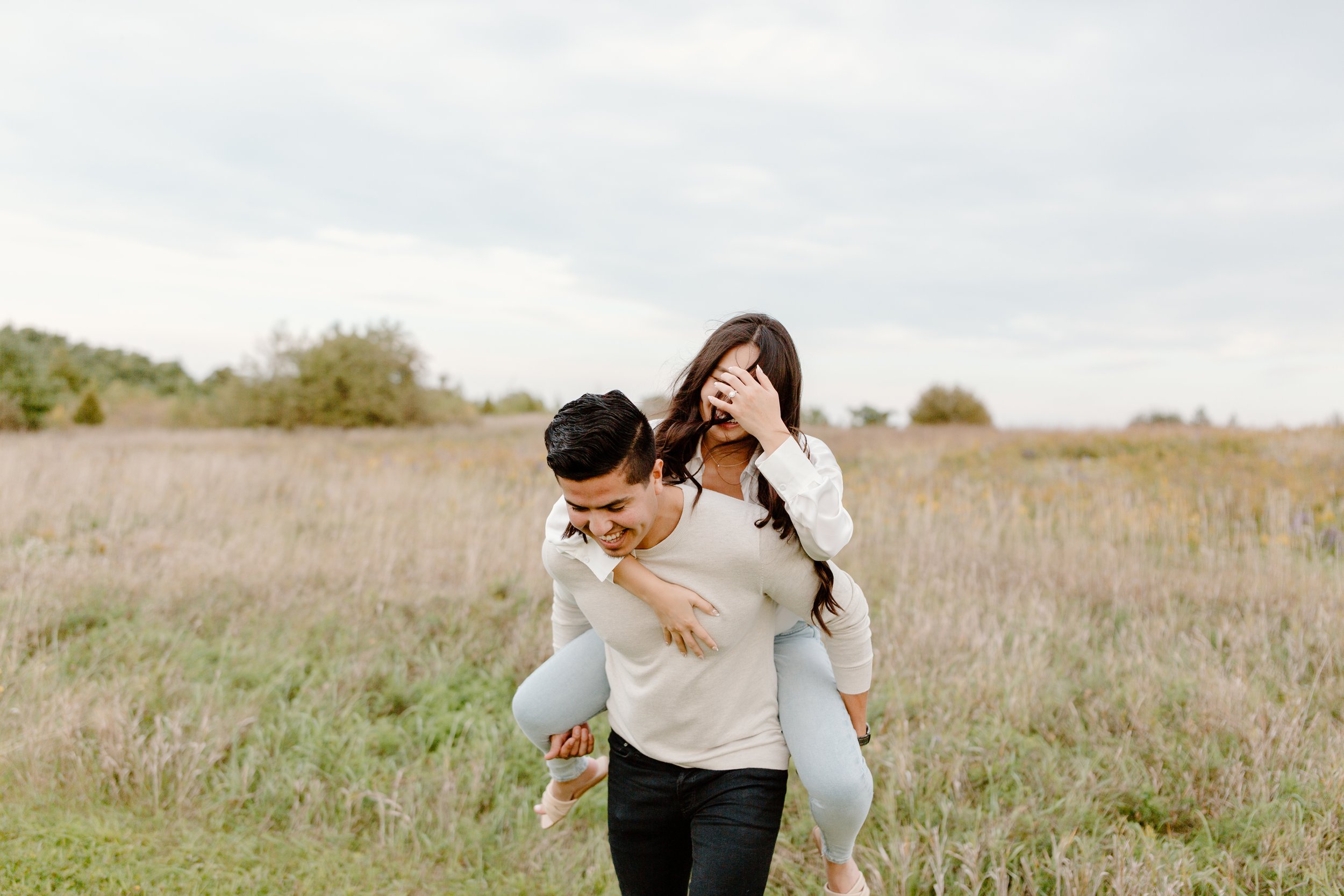 A young couple enjoying a playful moment outdoors in a field, with the woman on the man's back, both smiling and laughing.