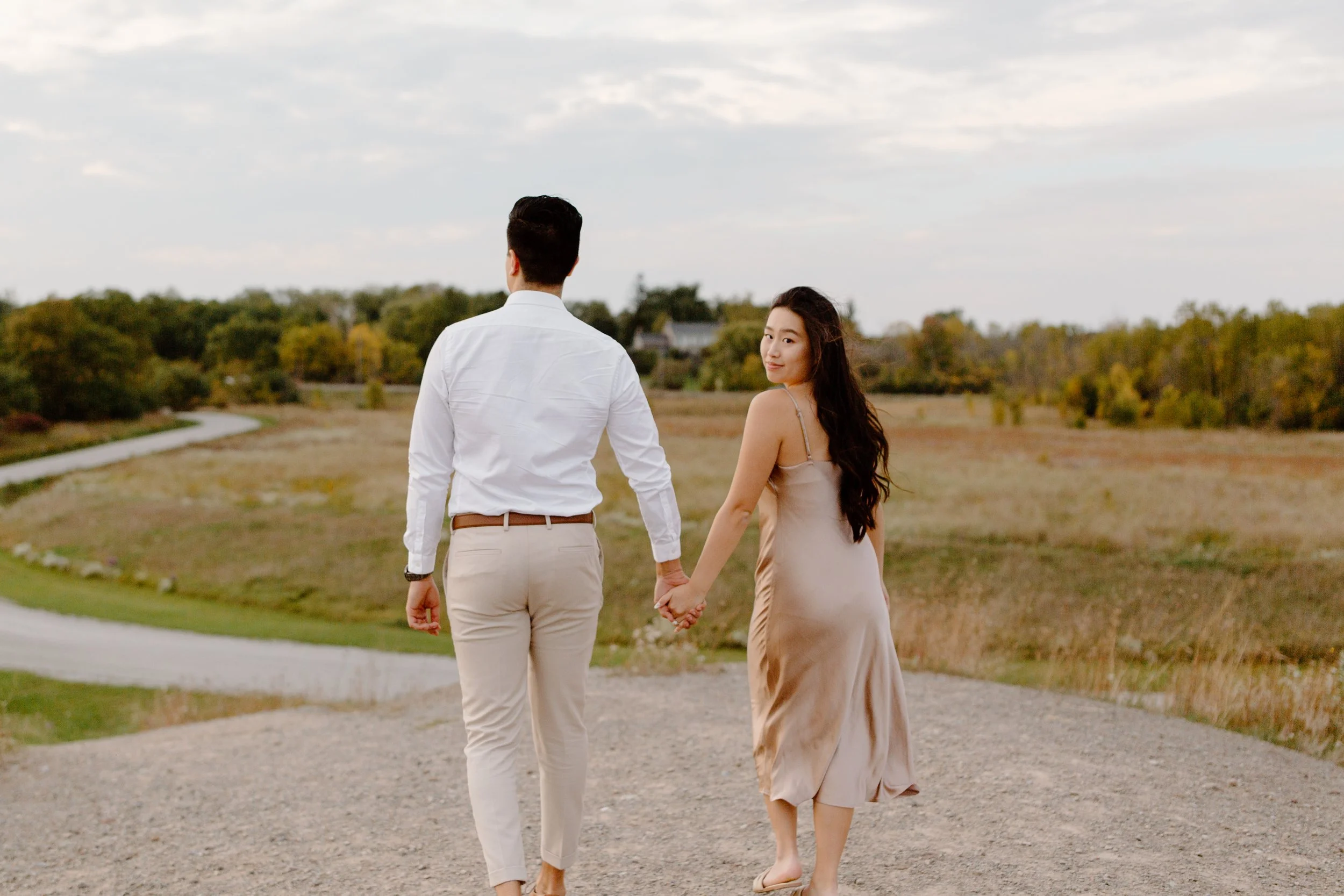 A young couple holding hands and walking on a gravel path in an open rural landscape with trees and a cloudy sky in the background.