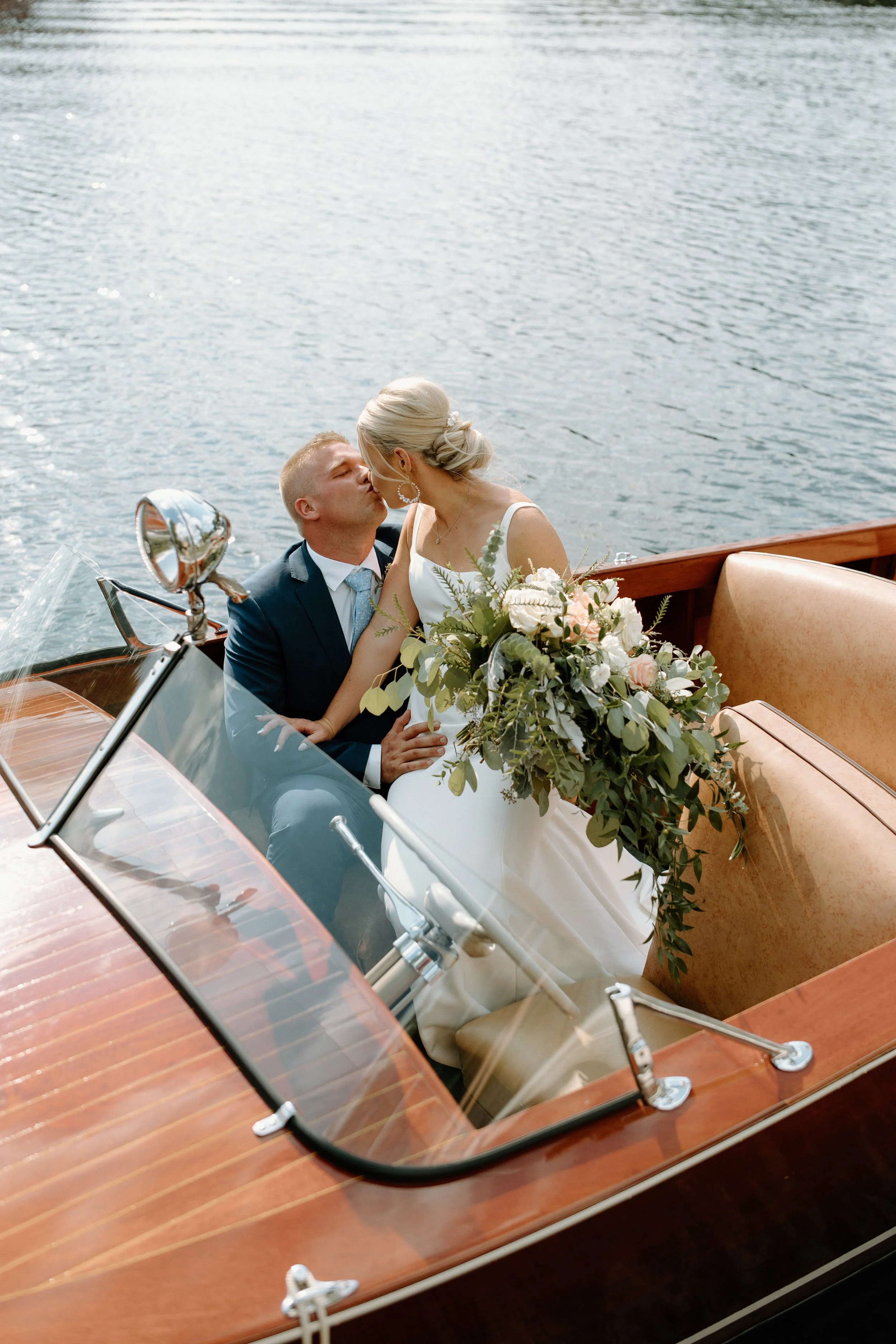 A newlywed couple sharing a kiss on a boat during sunset, with the bride holding a large bouquet of flowers and water in the background.