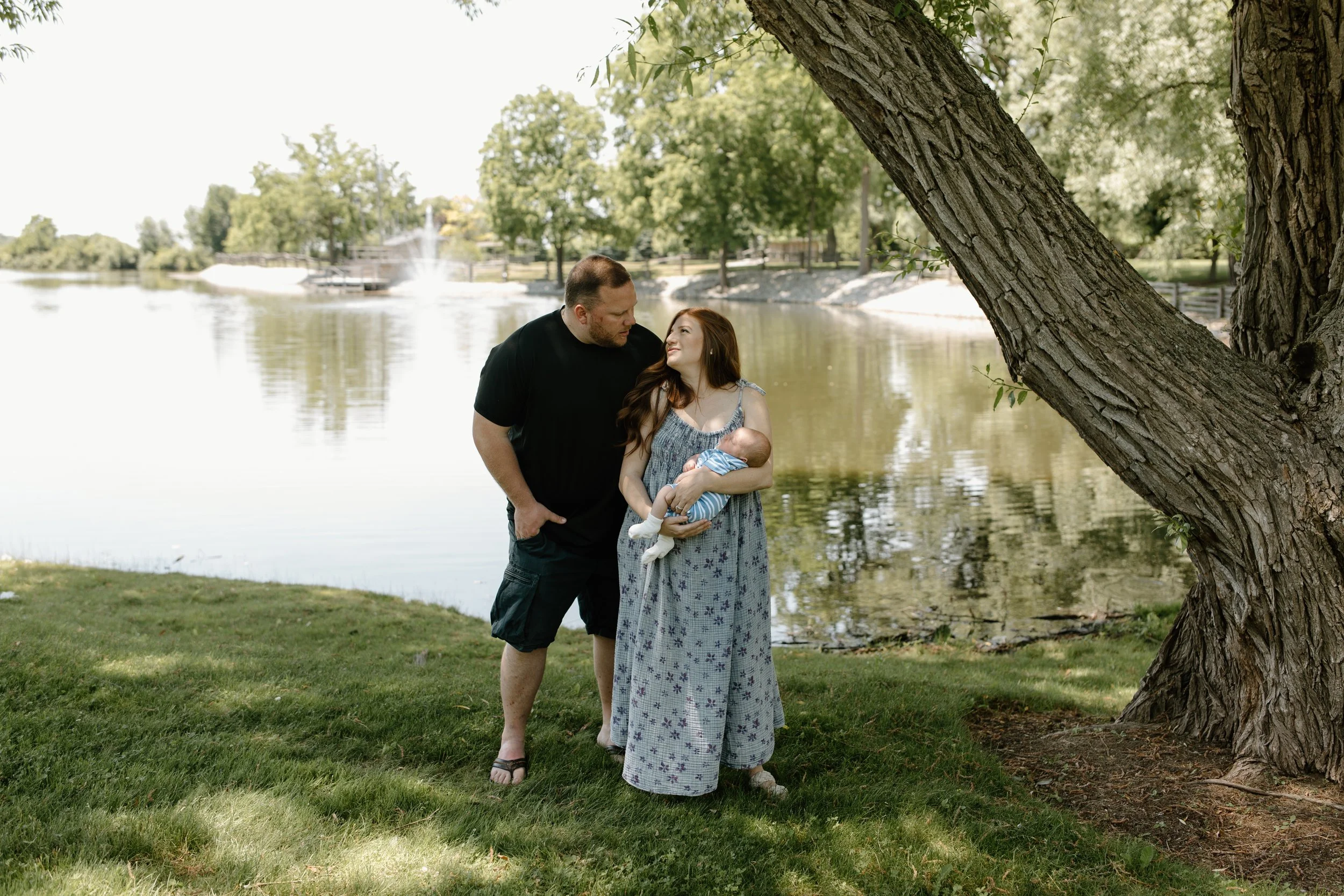 A family by a lake, with a woman holding a baby and a man looking at her, standing under a large tree in a park.