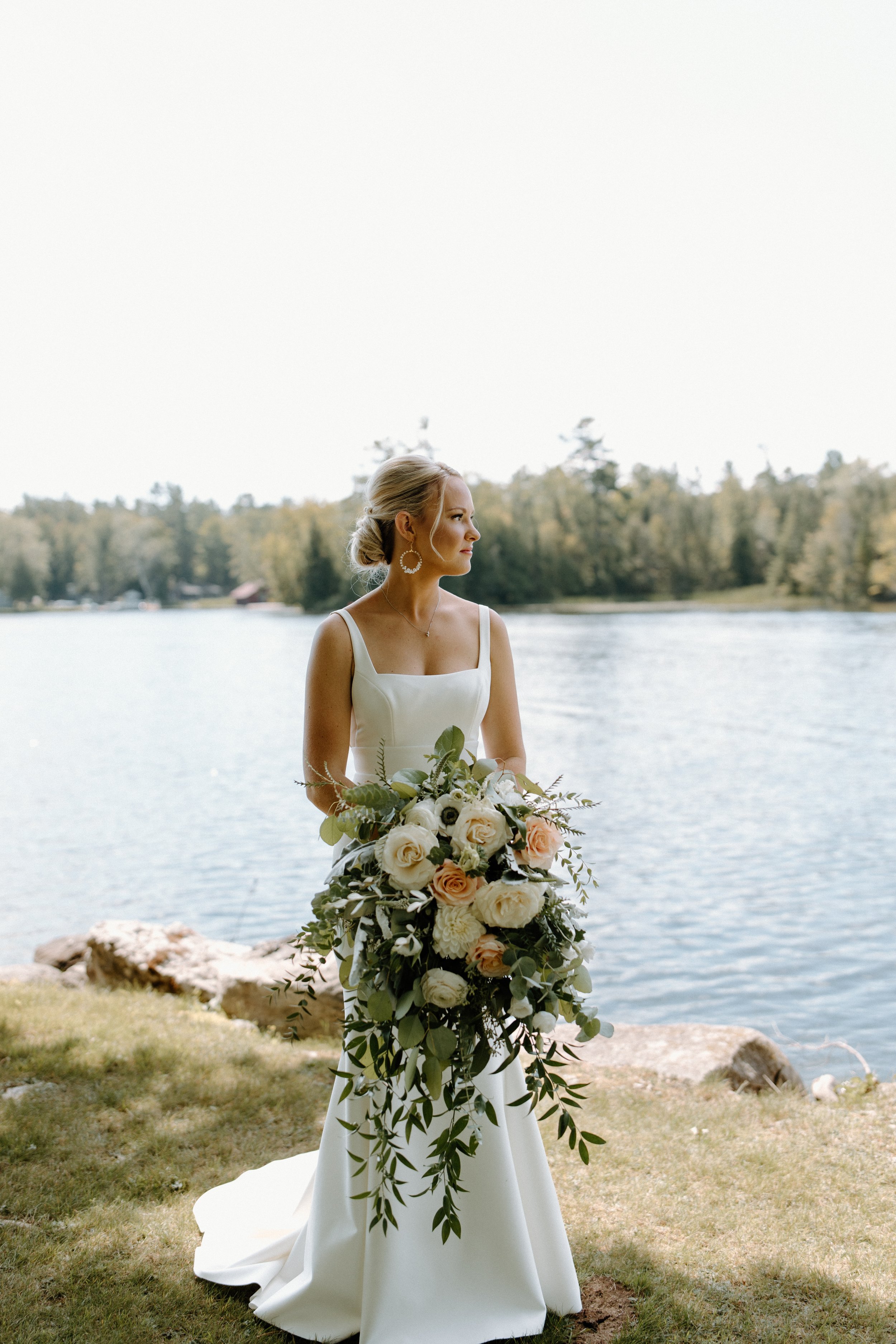 A bride holding a large bouquet of white and blush roses, greenery, and other flowers near a lakeside with trees in the background.